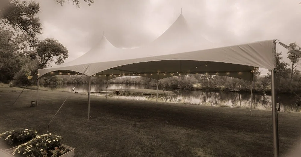 A large white event tent set up on a grassy area near a lake, with trees in the background and string lights hanging underneath the tent.
