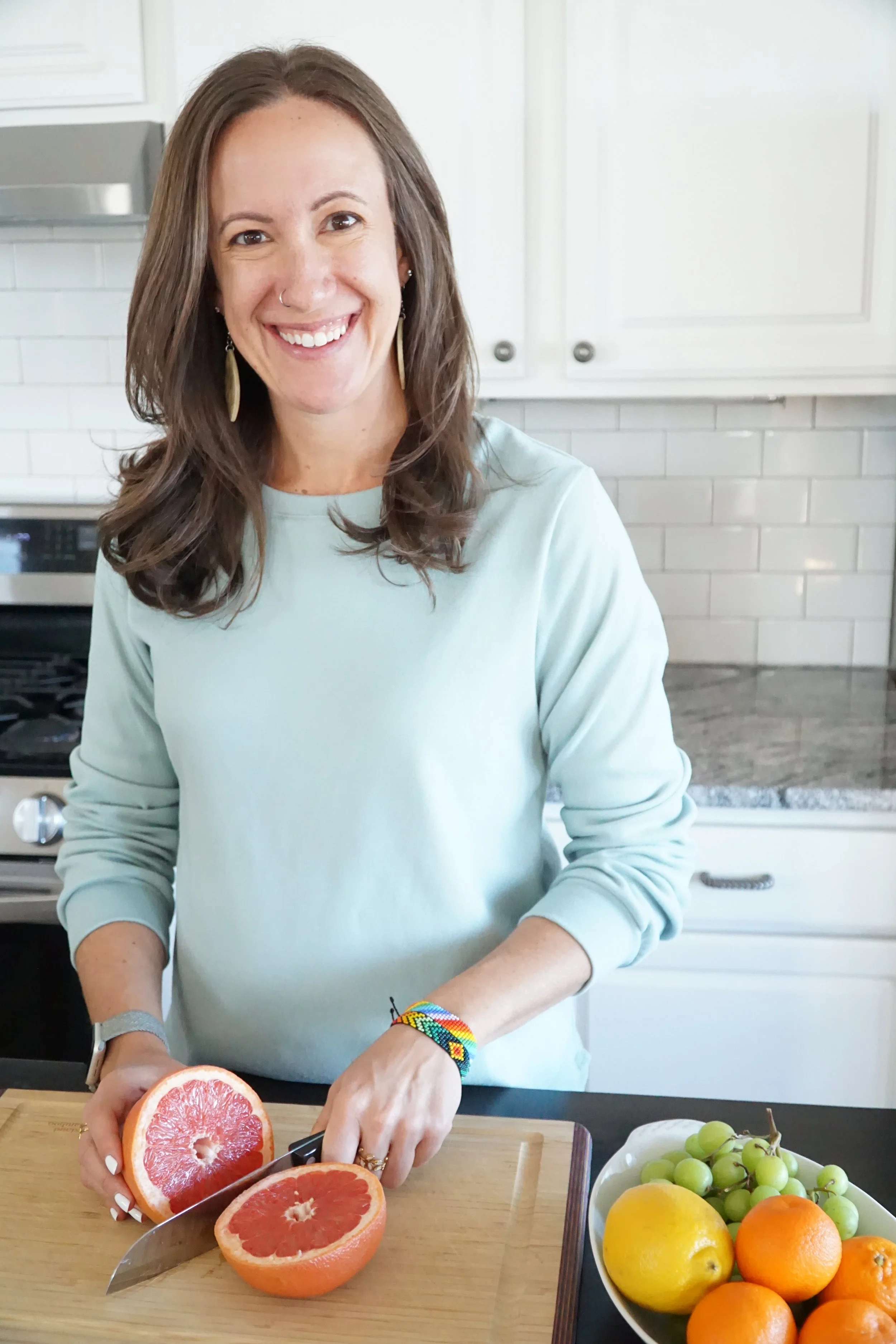 A woman smiling in a kitchen, slicing a grapefruit on a wooden cutting board. There is a bowl of assorted fruits including green grapes, oranges, and lemons on the table.