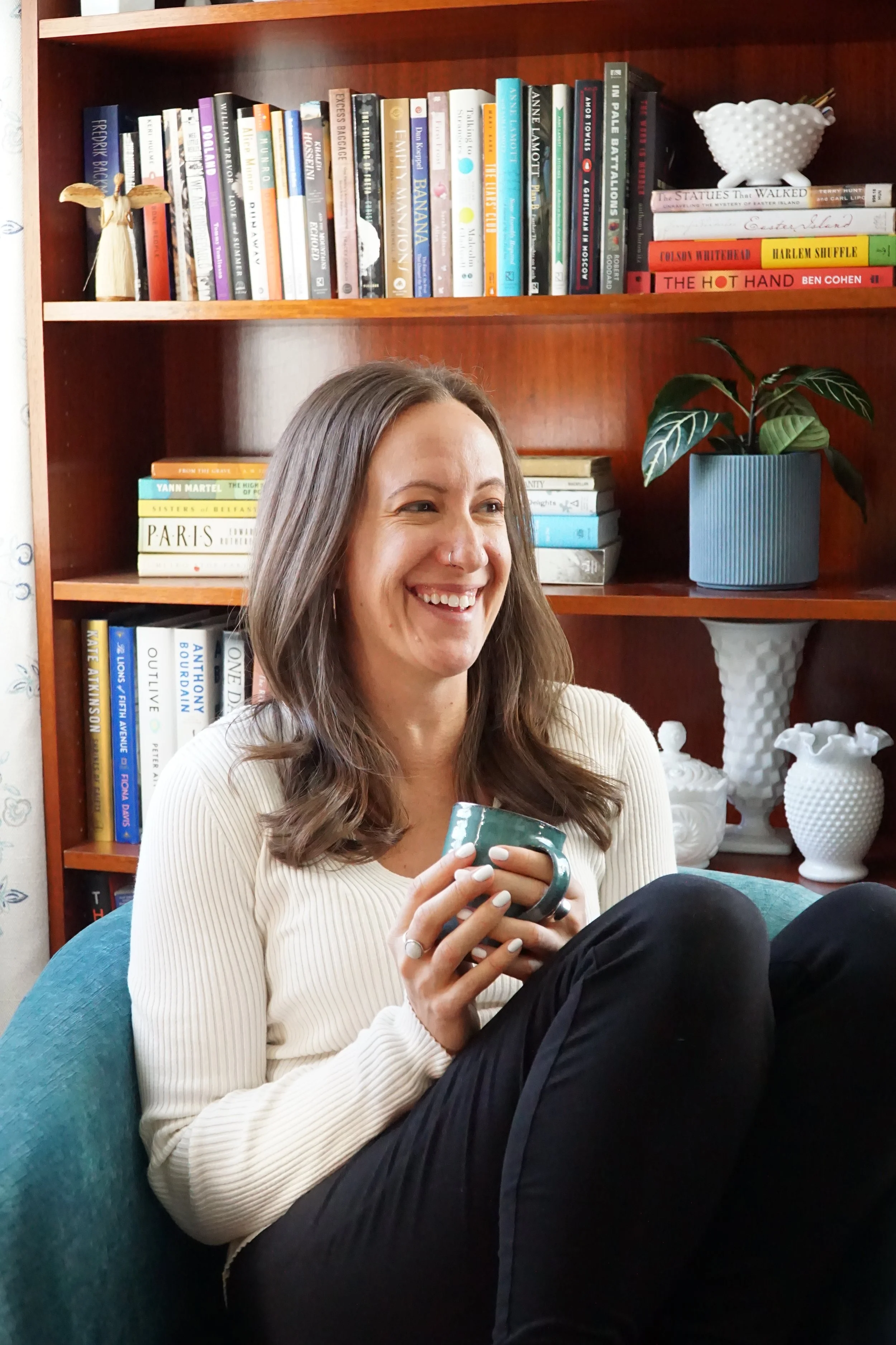 A woman with long brown hair, smiling, sitting on a teal chair while holding a ceramic mug. She is wearing a cream ribbed sweater and black pants, with a bookshelf behind her filled with books, a potted plant, and decorative white vases.