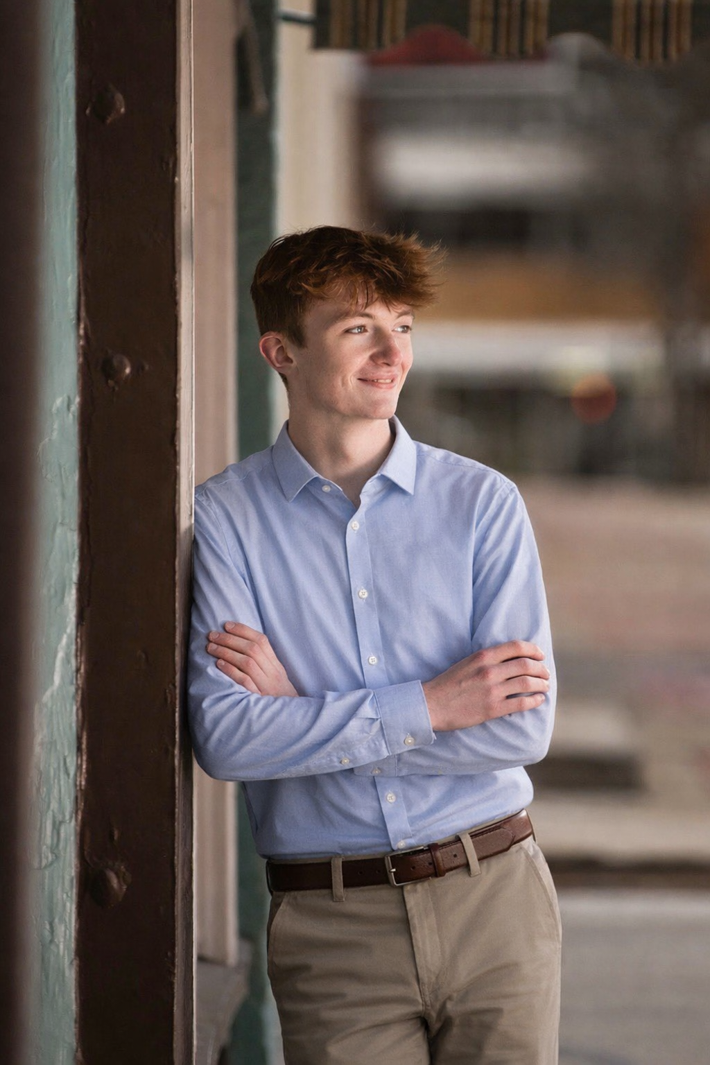 Young man with brown hair and light skin wearing a light blue button-down shirt and khaki pants, leaning against a wooden column with arms crossed, smiling and looking to the side in an indoor setting.
