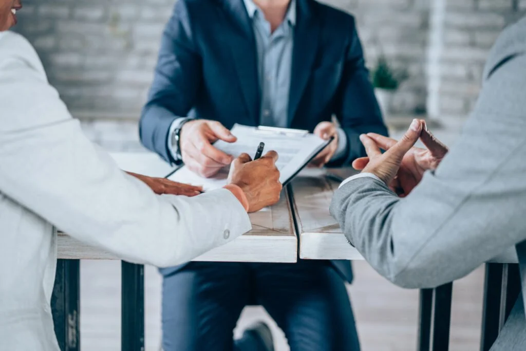 Three business professionals in a meeting, one taking notes on a clipboard, two others engaged in discussion, at a wooden conference table in an office setting.