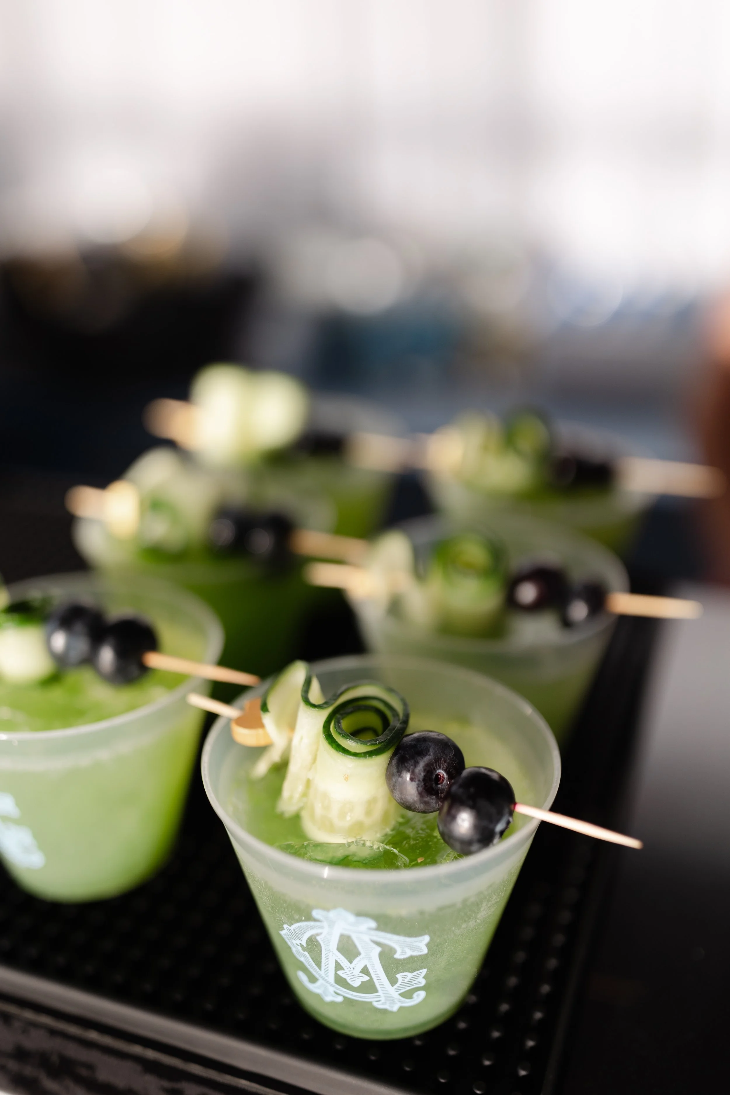 Green cocktails garnished with cucumber spirals and blueberries, served in clear cups with a logo.