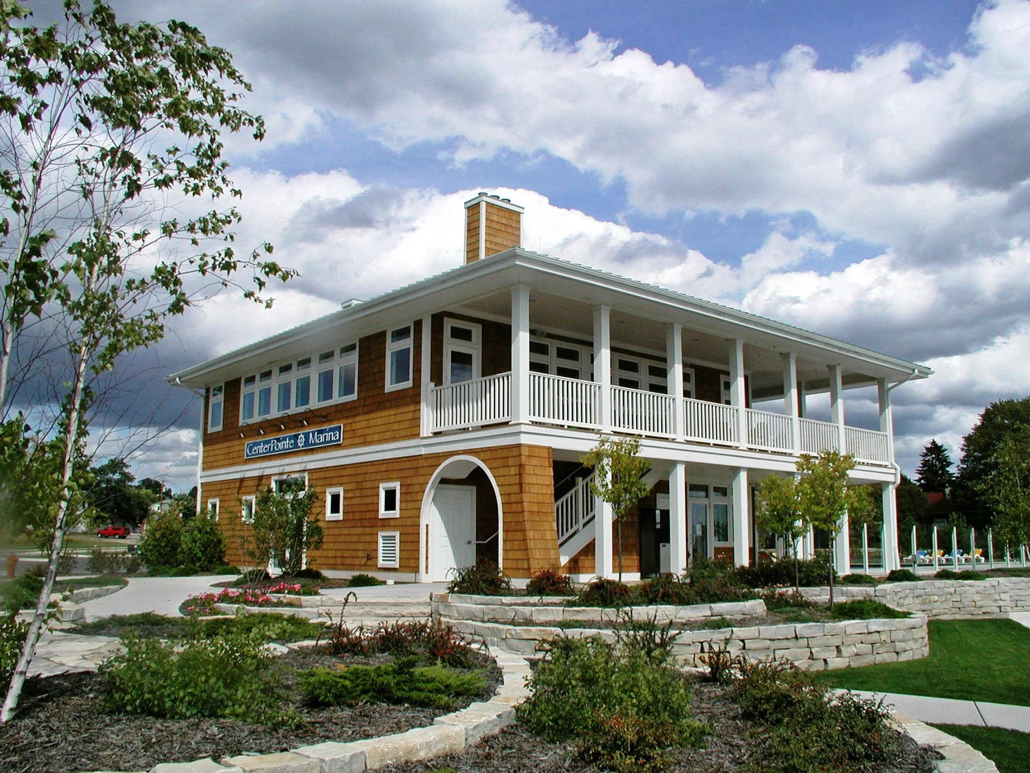 A two-story building with brown shingles and white trim, a balcony on the second floor, and landscaped garden in front, under a partly cloudy sky.