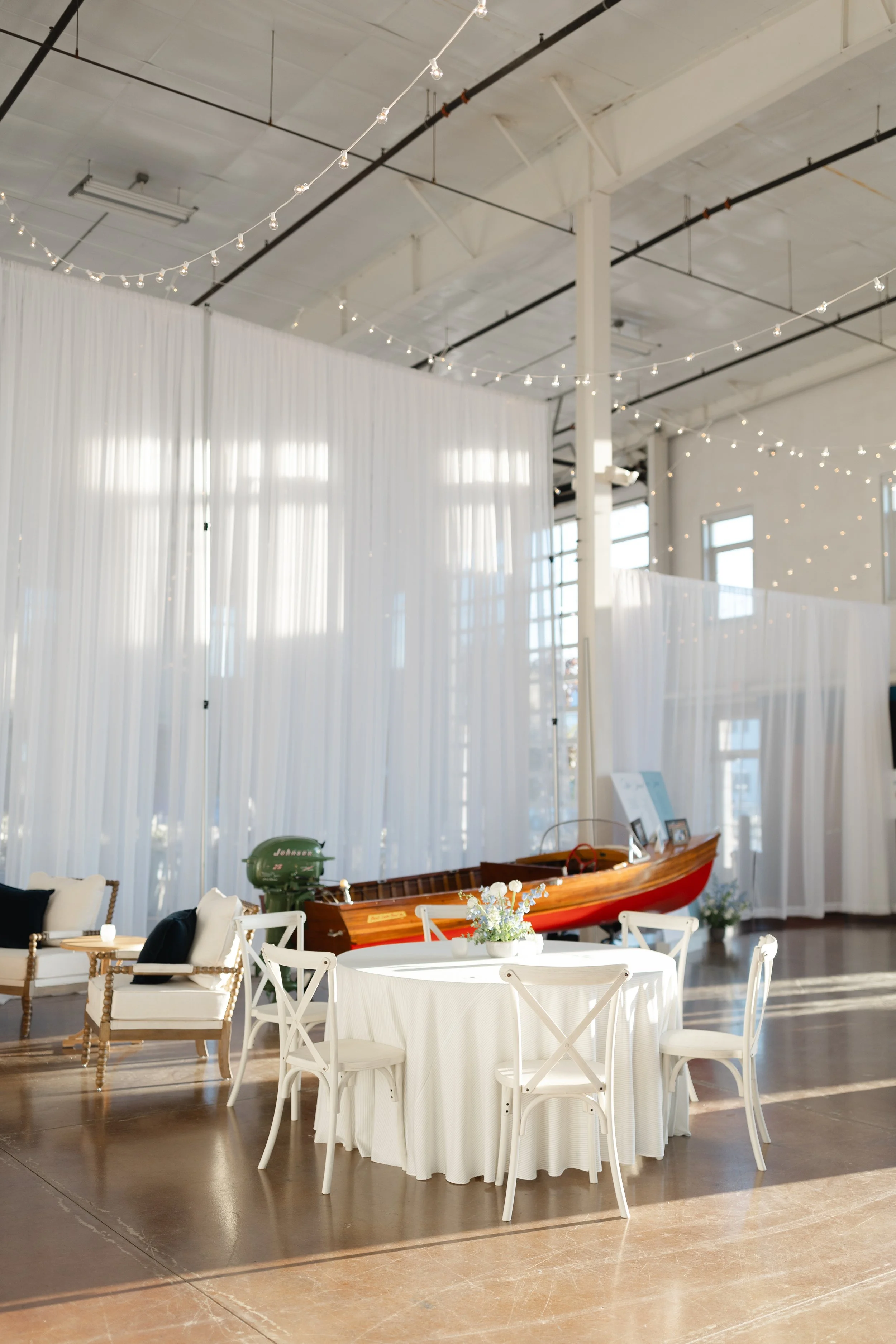 Indoor event space with white draped curtains, string lights, a round table with white tablecloth and flower centerpiece, white chairs, and a wooden boat on display.