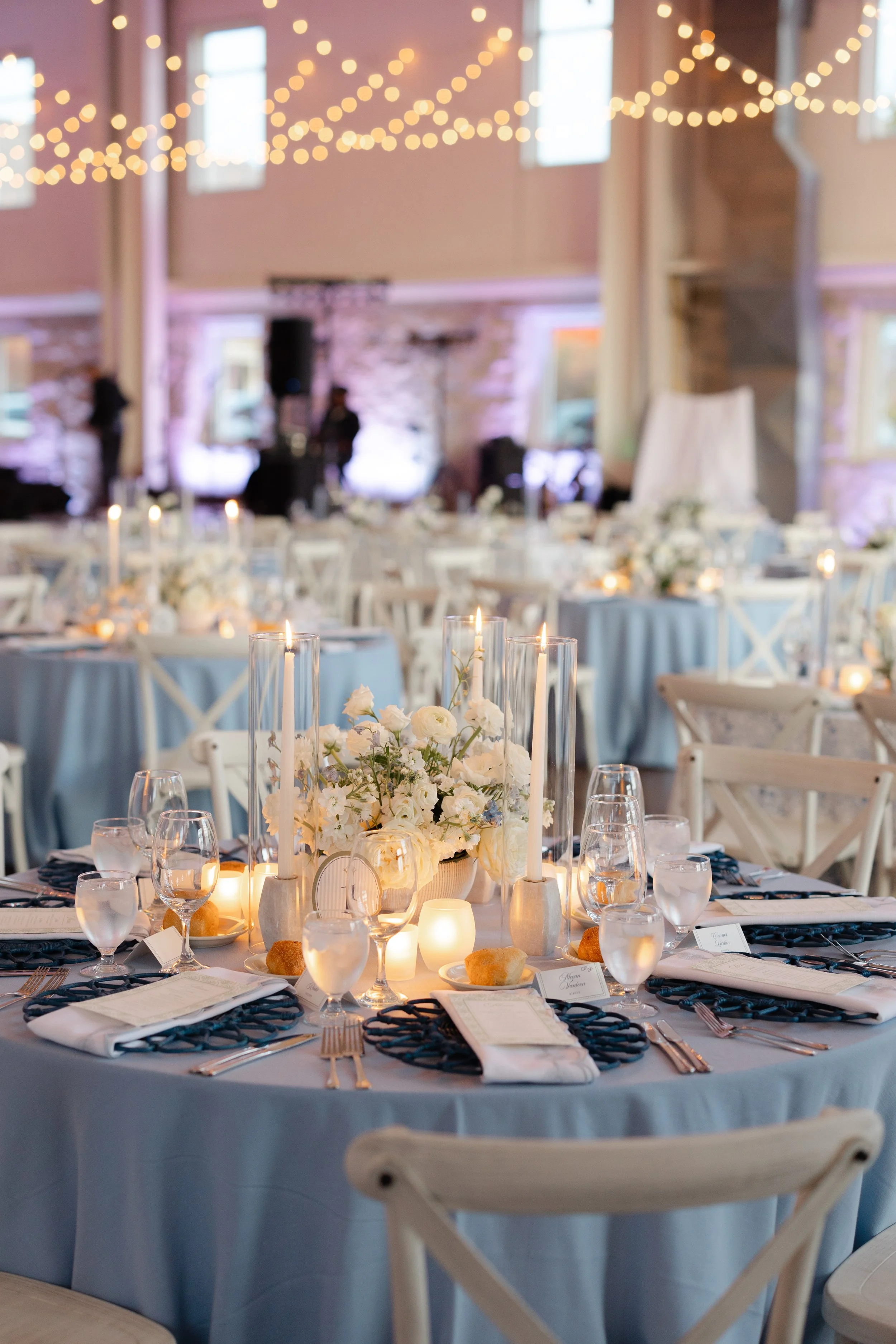 Elegant indoor wedding reception with round tables decorated with white flowers, candles, and navy blue napkins, set with glassware and silverware, with string lights hanging from the ceiling in a rustic venue with stone walls.