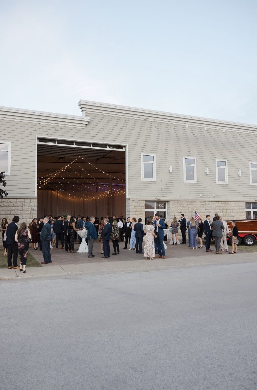 People dressed in formal attire gather outside a large building, possibly for a wedding or celebration, with string lights hanging inside.