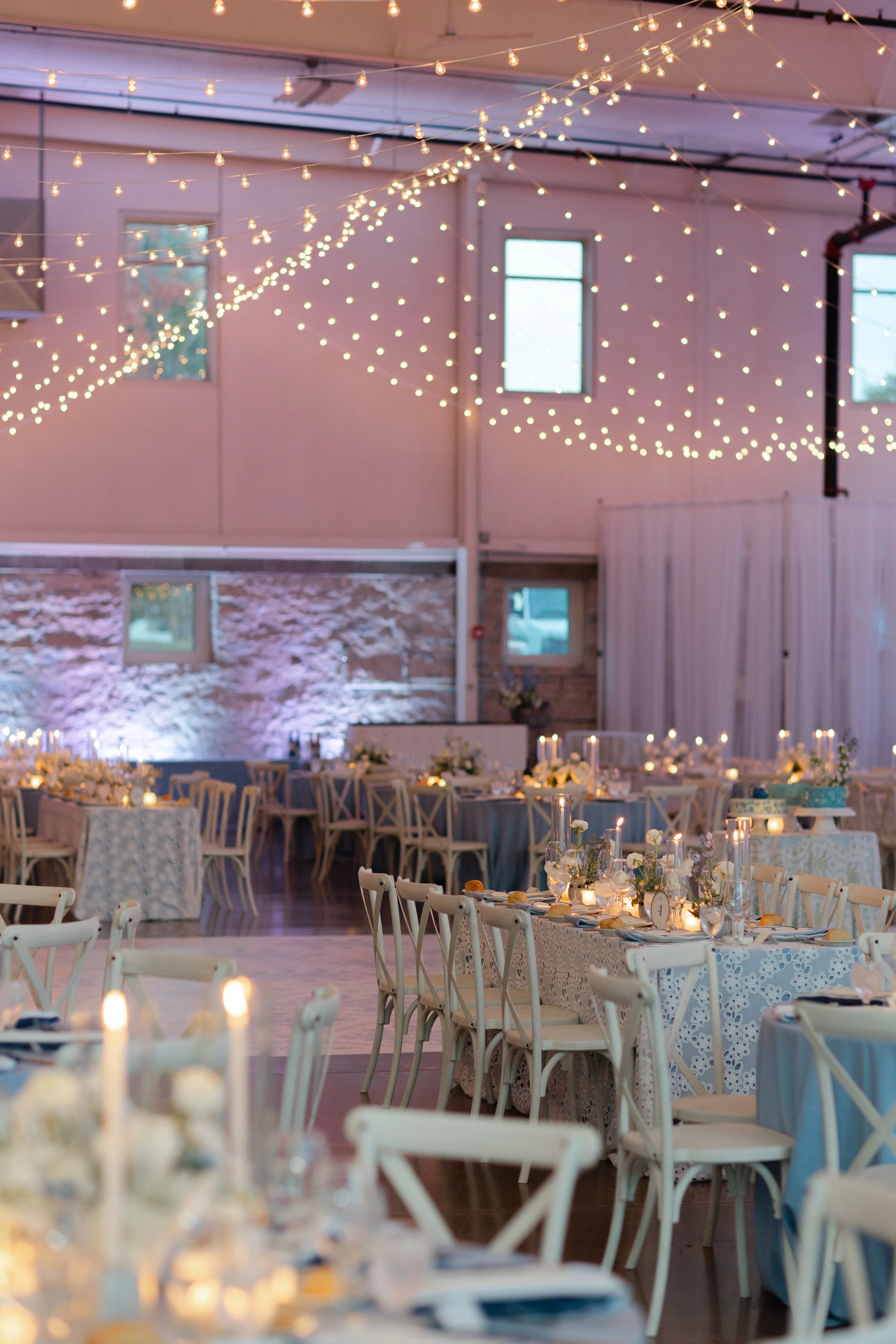 Wedding reception decorated with string lights hanging from the ceiling, elegant tables with white chairs, floral centerpieces, and candles, set in a spacious venue with a rustic stone wall and large windows.