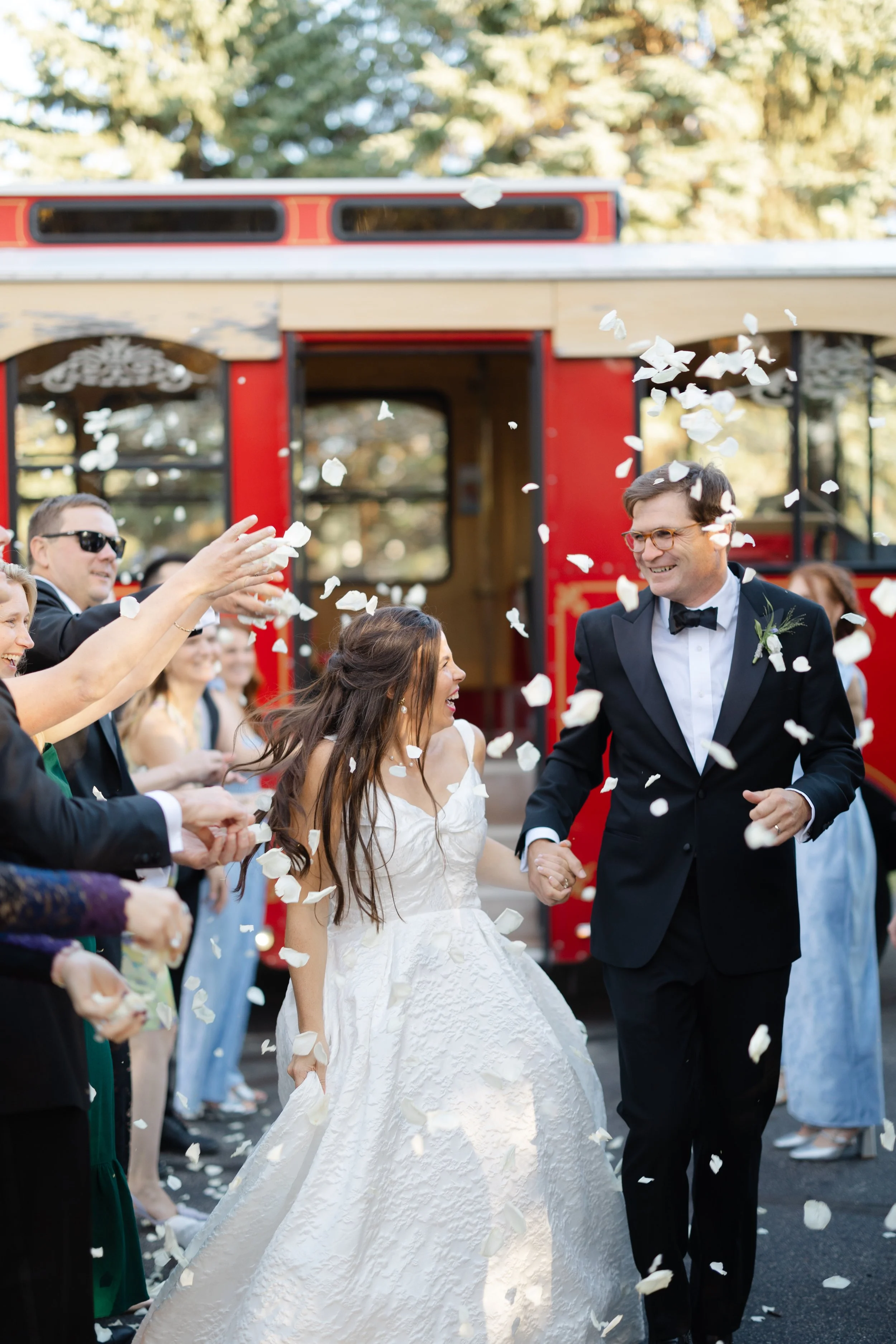 A newlywed couple celebrating their wedding day with guests throwing white flower petals in front of a red trolley car during daytime.