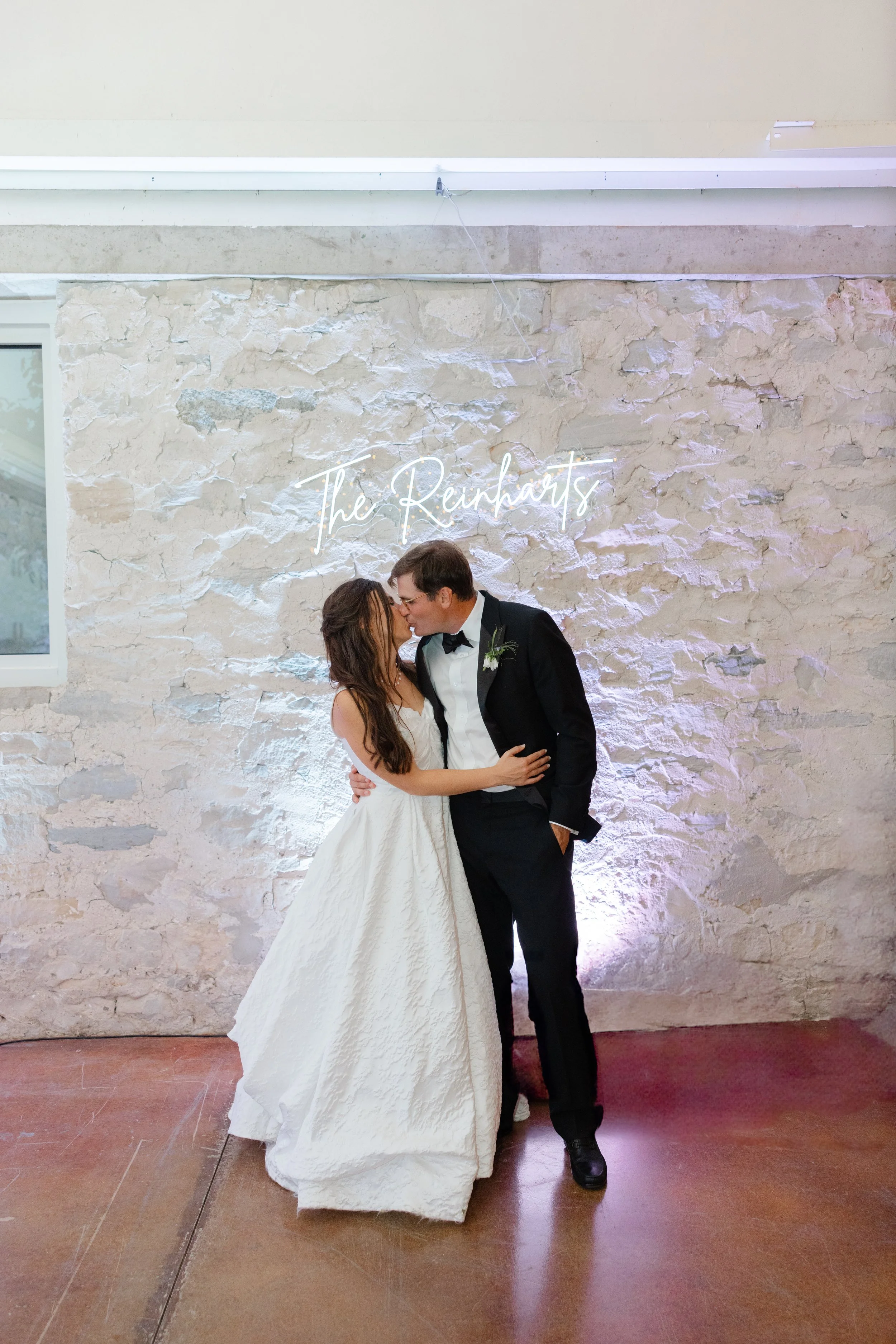 A bride and groom kissing at their wedding reception in front of a white brick wall with a neon sign reading 'The Reunions'.