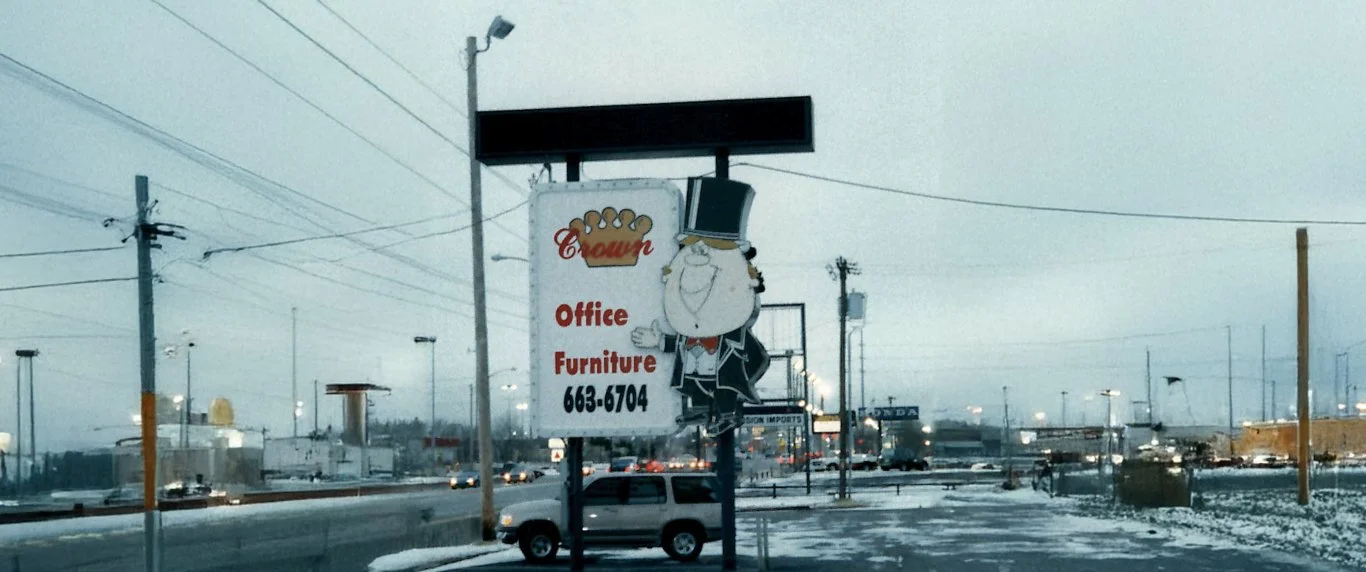 A street scene with a sign for Crown office furniture, featuring a cartoon character of a loaf of bread with a face, dressed in a tuxedo, holding a top hat. The background shows a snowy day with traffic and power lines.