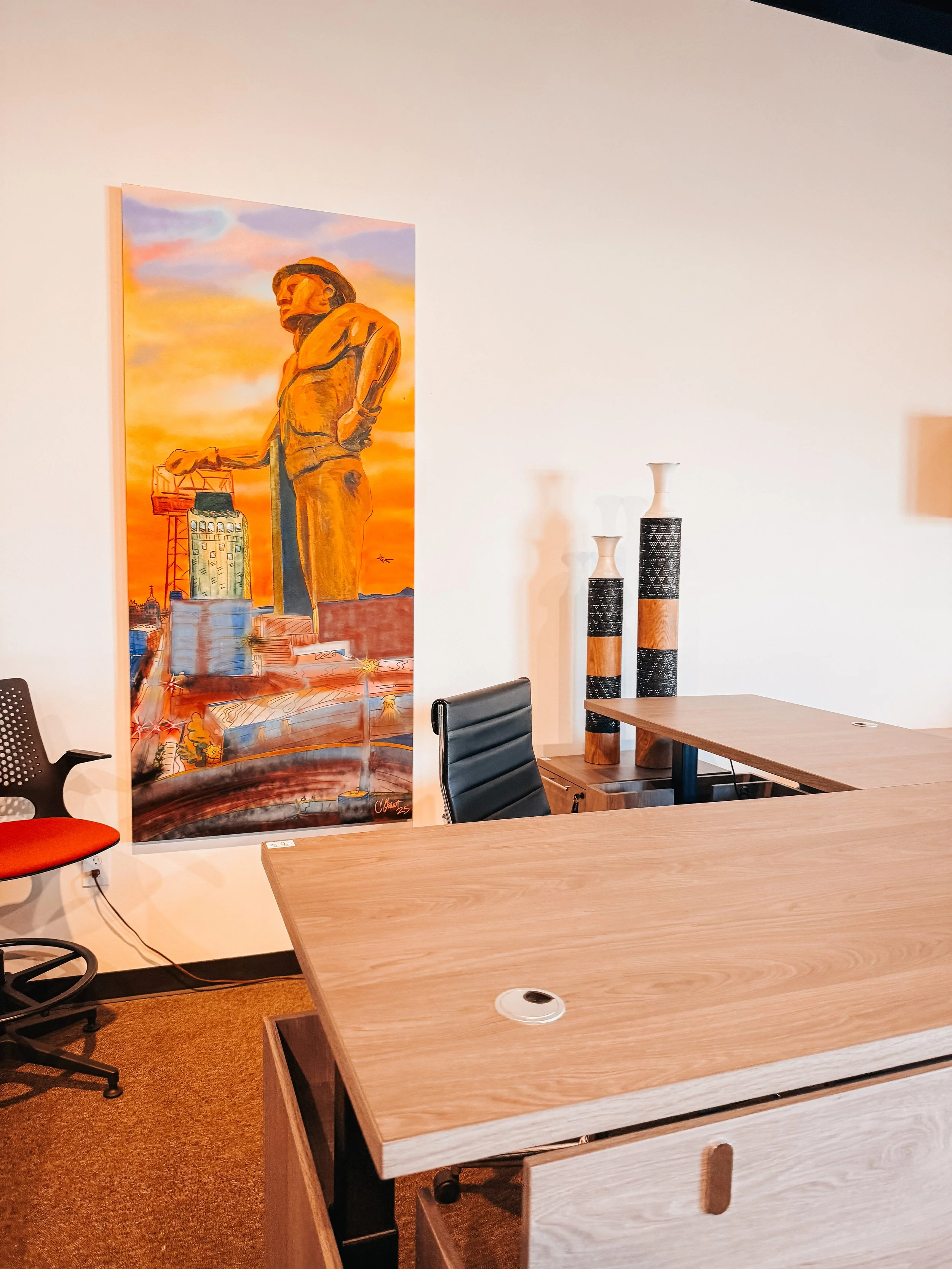 Office workspace with L-shaped wood-grain desk, black padded executive chair, cable management grommet, decorative wooden vases, and colorful cityscape painting