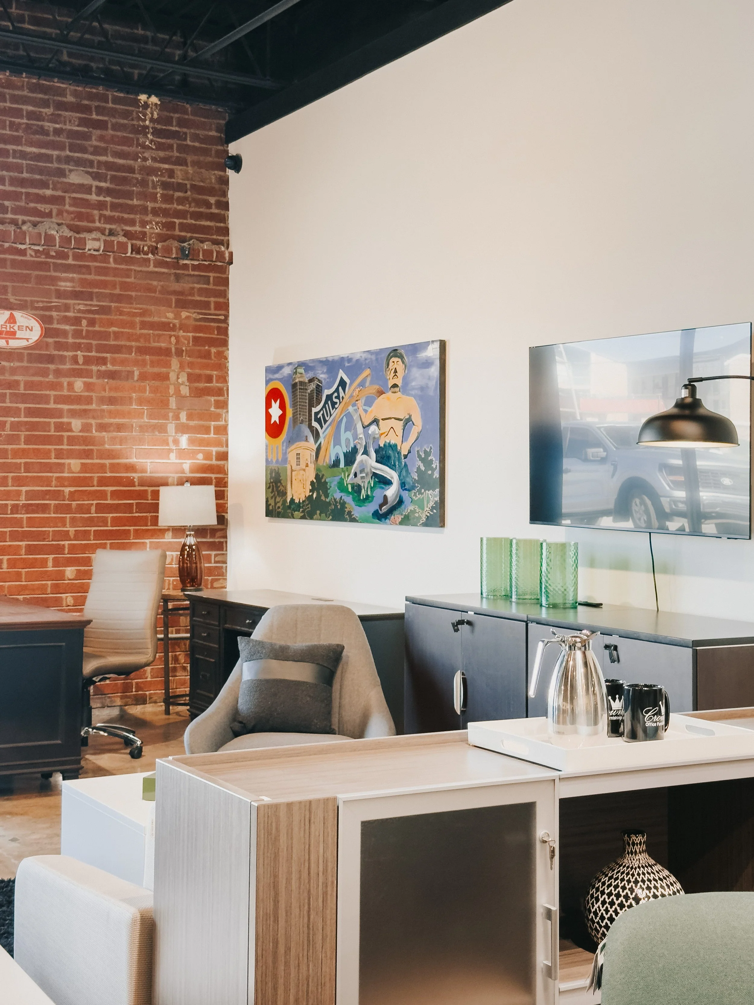 Crown Office Furnishings showroom featuring executive desk, tan ribbed office chair, gray lounge chair, Tulsa Oklahoma artwork, and industrial exposed brick wall⁠
