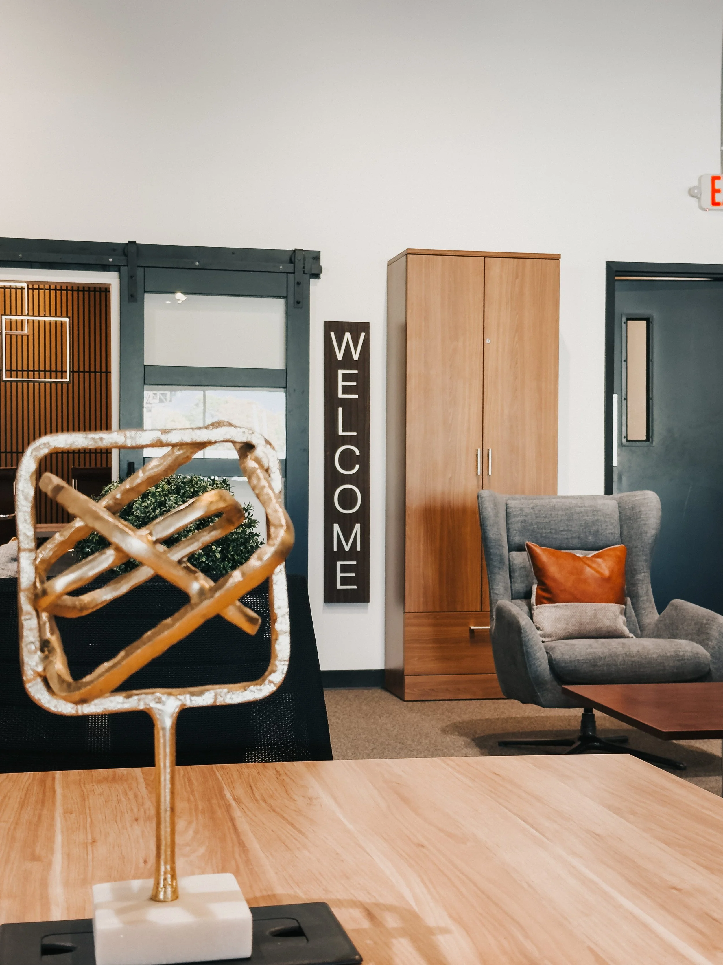 Crown Office Furnishings showroom lobby with gray wingback armchair, wood storage wardrobe, sliding glass barn door, and gold desk sculpture