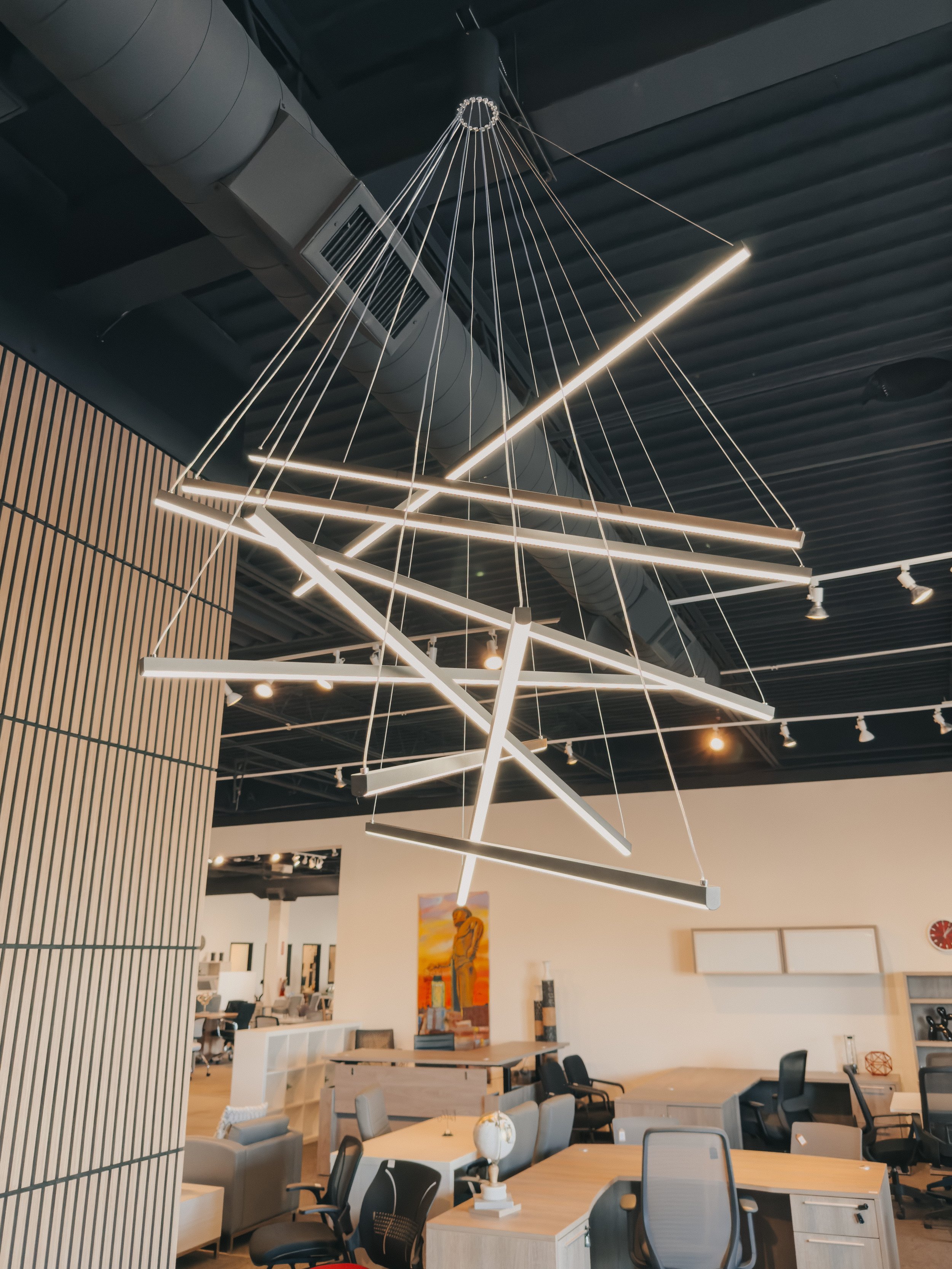 Crown Office Furnishings showroom floor displaying wood desks, ergonomic office chairs, storage cabinets, and modern geometric LED chandelier under industrial ceiling⁠