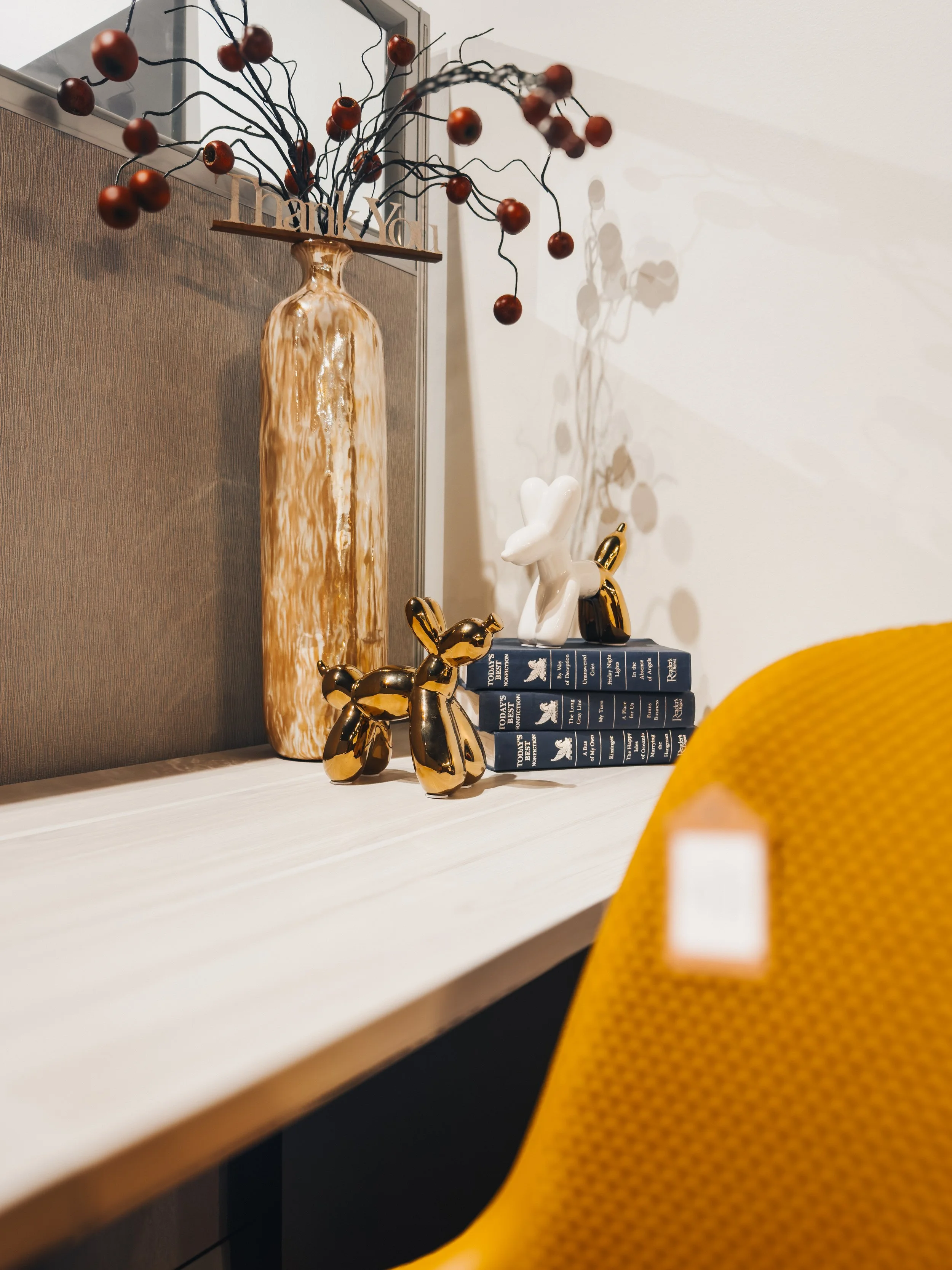 Gold vase with decorative berry branches, metallic balloon dog figurines, and stacked books styled on a wood-grain office desk with yellow perforated accent chair