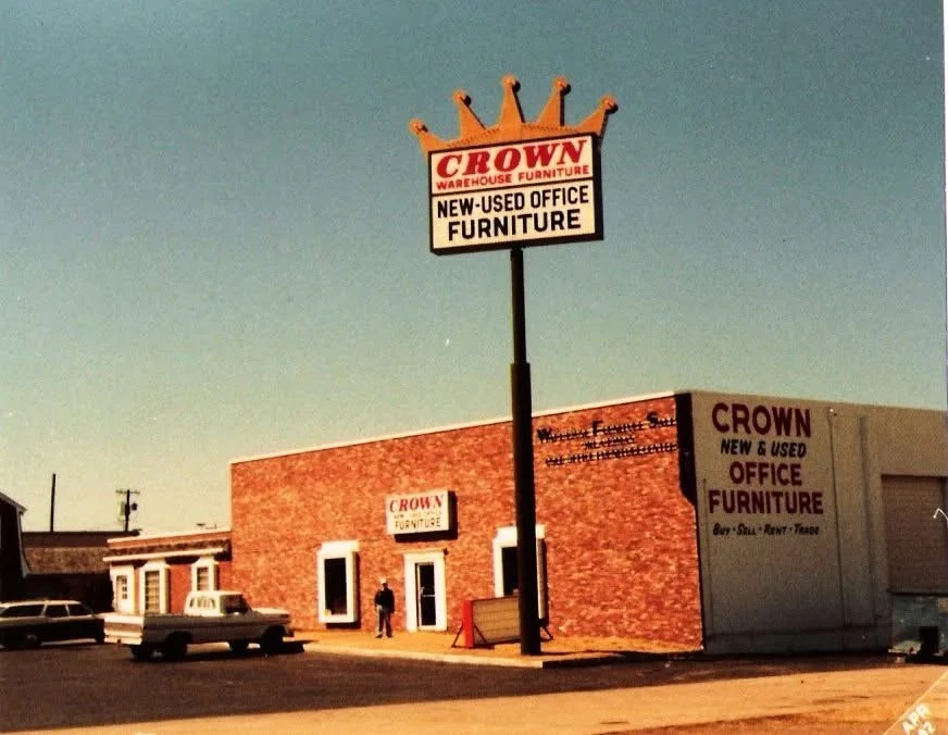A brick building with signs for Crown Warehouse Furniture and a tall sign with a crown logo advertising new and used office furniture.