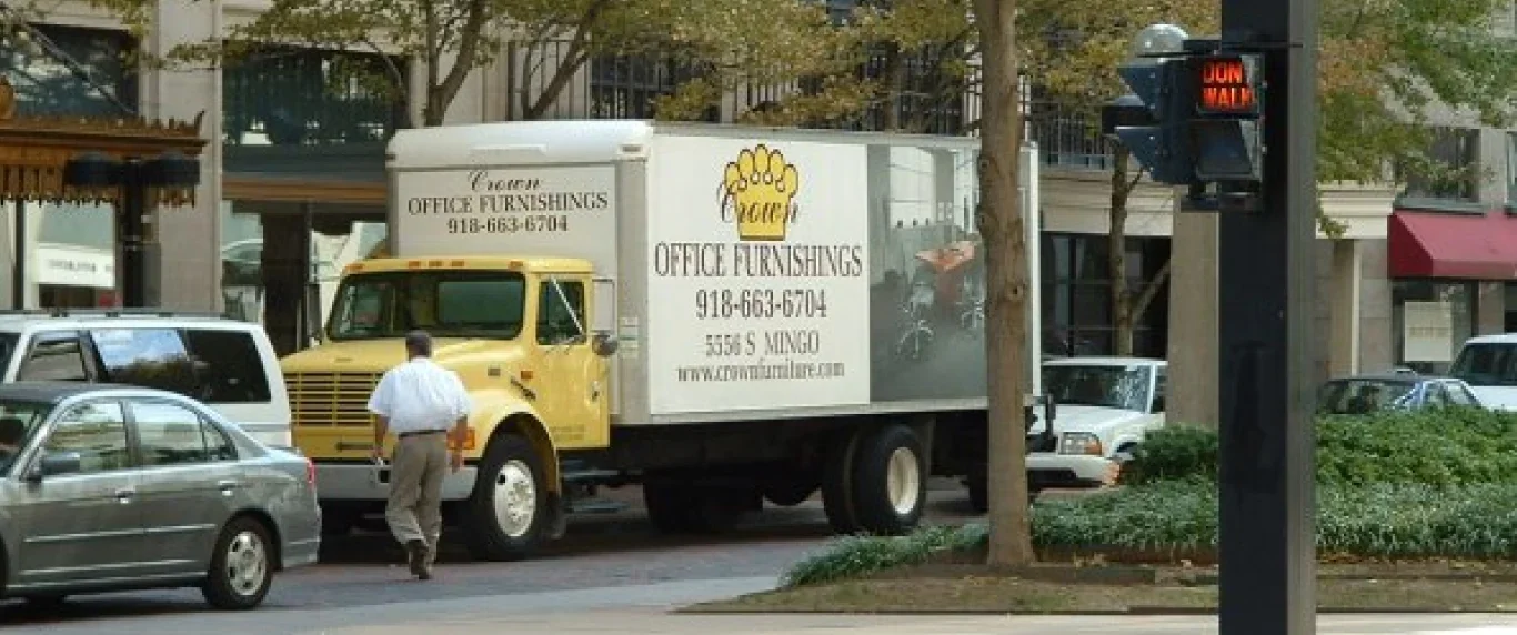 A yellow box truck with an ad for office furnishings parked on a city street. There are several cars around, and a man in a white shirt and khaki pants walking nearby. A traffic light displays a "DON'T WALK" sign.