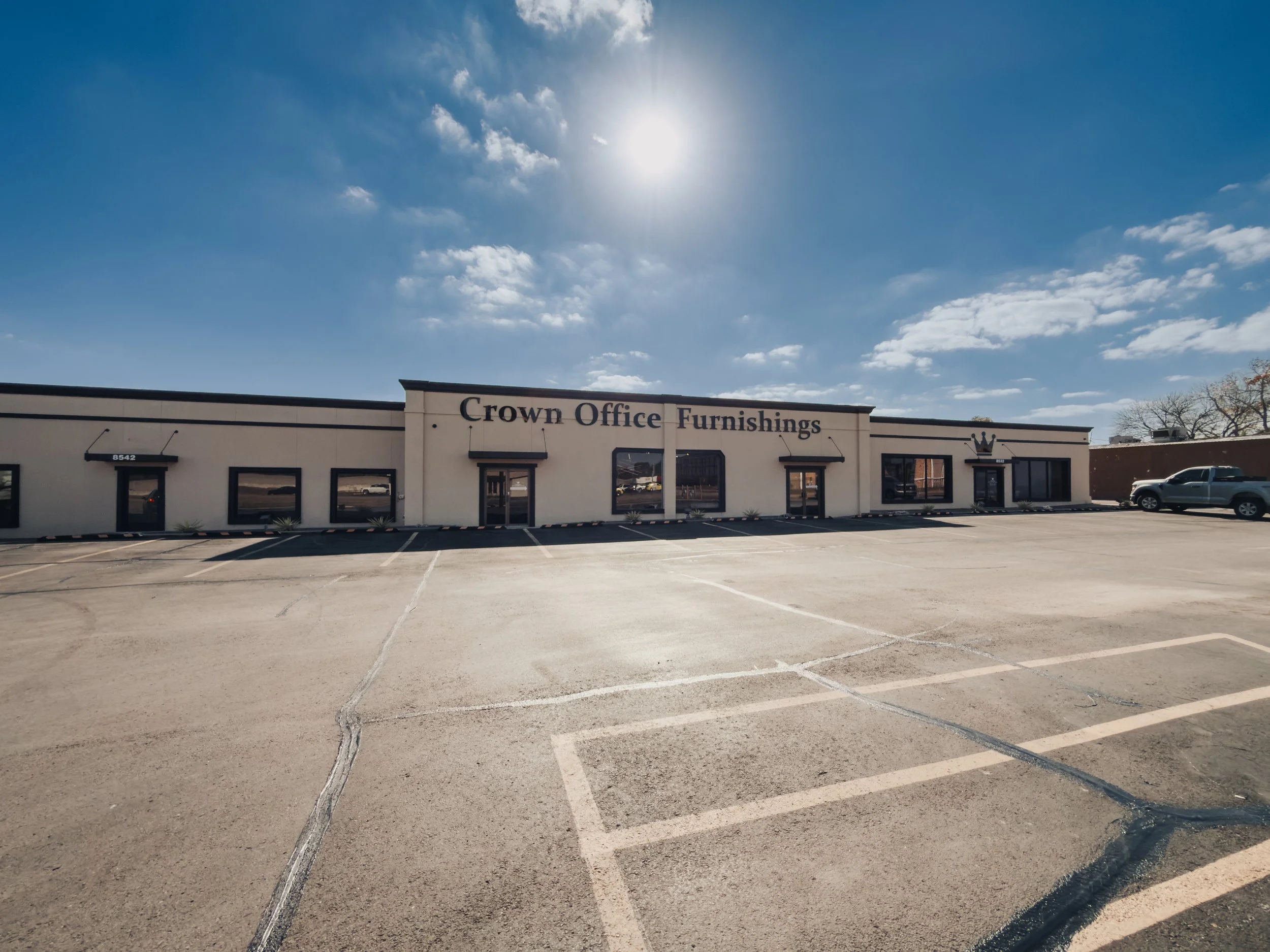 Empty parking lot in front of a building with the sign 'Crown Office Furnishings' under a bright, sunny sky with some scattered clouds.