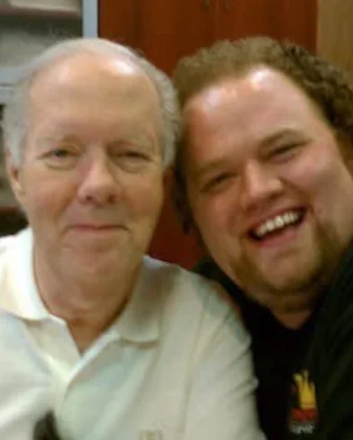Two men are smiling and taking a selfie together. The older man on the left has gray hair and is wearing a white polo shirt. The younger man on the right has curly hair and is wearing a dark shirt.