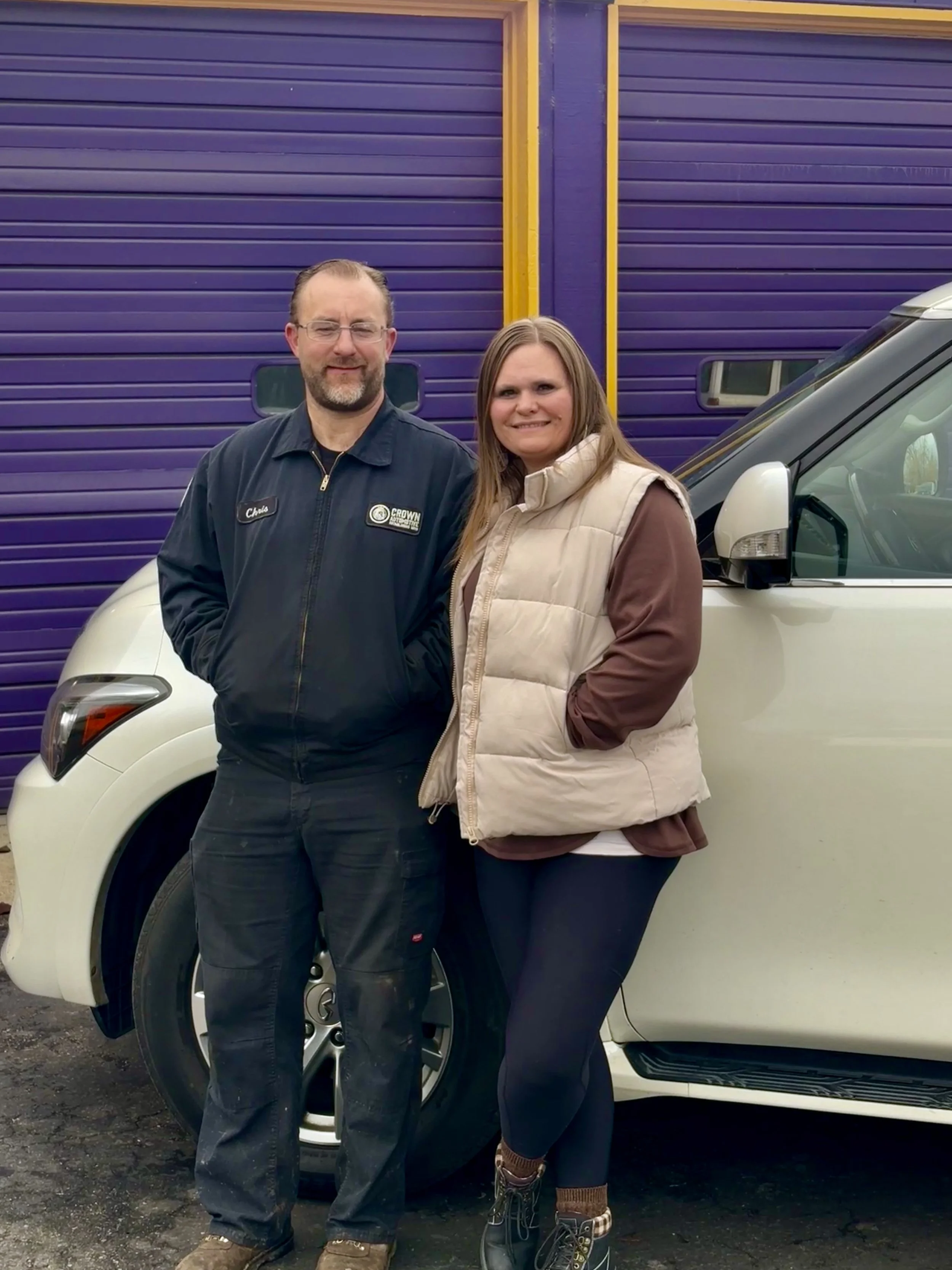 A man and a woman stand side by side in front of a white vehicle with a purple and yellow background, smiling at the camera.