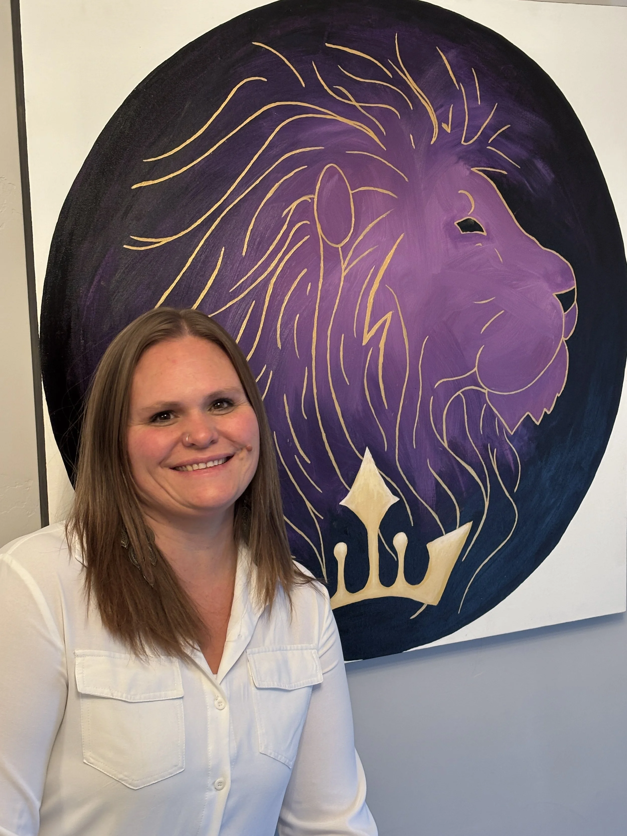 A woman with shoulder-length brown hair, wearing a white shirt, smiling in front of a large mural of a purple lion with a gold outline and gold crown.