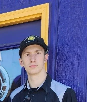 A young man wearing a black cap with a logo, standing against a purple wall with yellow trim.