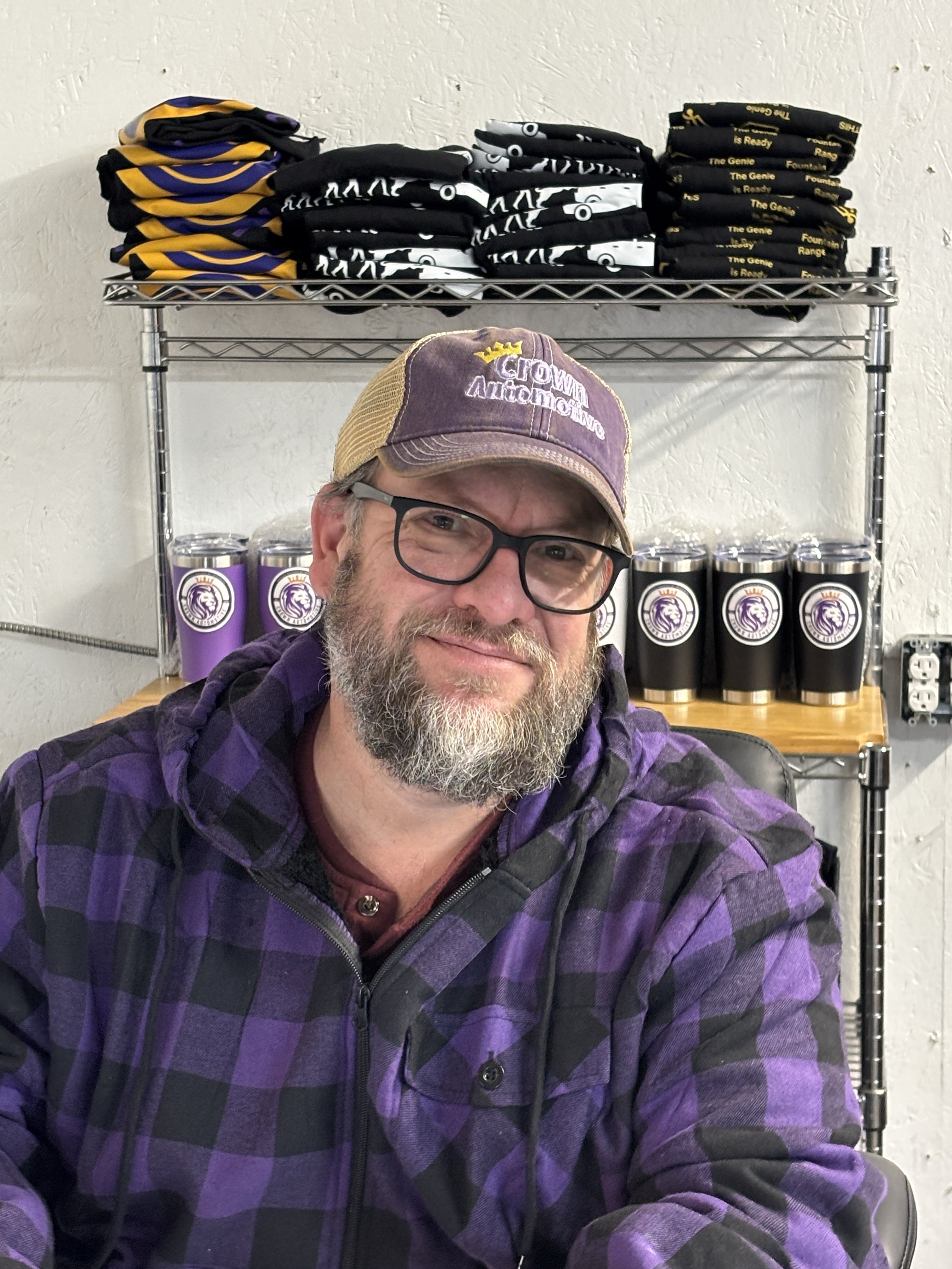 A man with glasses and a beard wearing a purple cap and purple checkered hoodie, smiling, sitting in front of shelves with purple and black tumbler cups and folded T-shirts in black, yellow, and white colors.