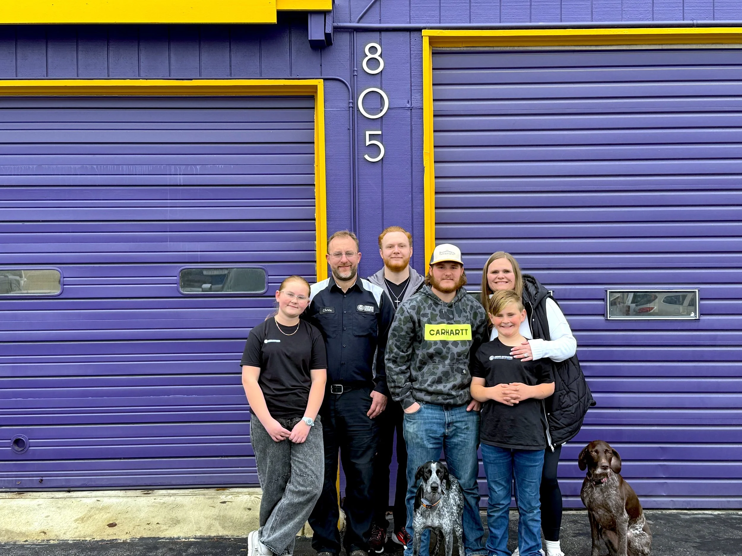 A group of six people and two dogs pose in front of a purple building with yellow accents and the number 805 on the wall.