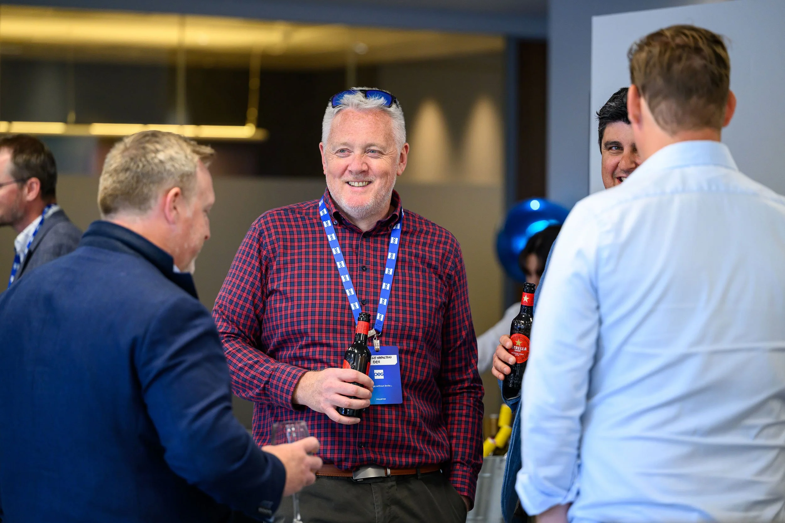 Four men at a professional networking event, holding drinks and chatting, with one man smiling and wearing sunglasses on his head.