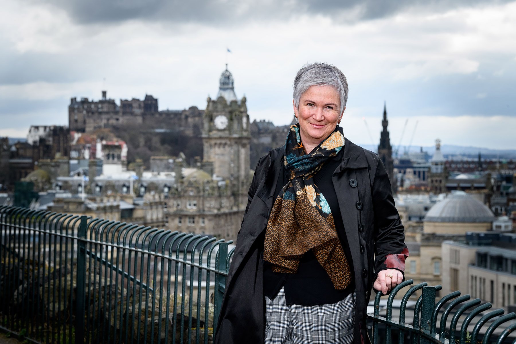 A smiling woman with short gray hair stands on a rooftop overlooking Edinburgh, Scotland, with Edinburgh Castle and historical buildings in the background.