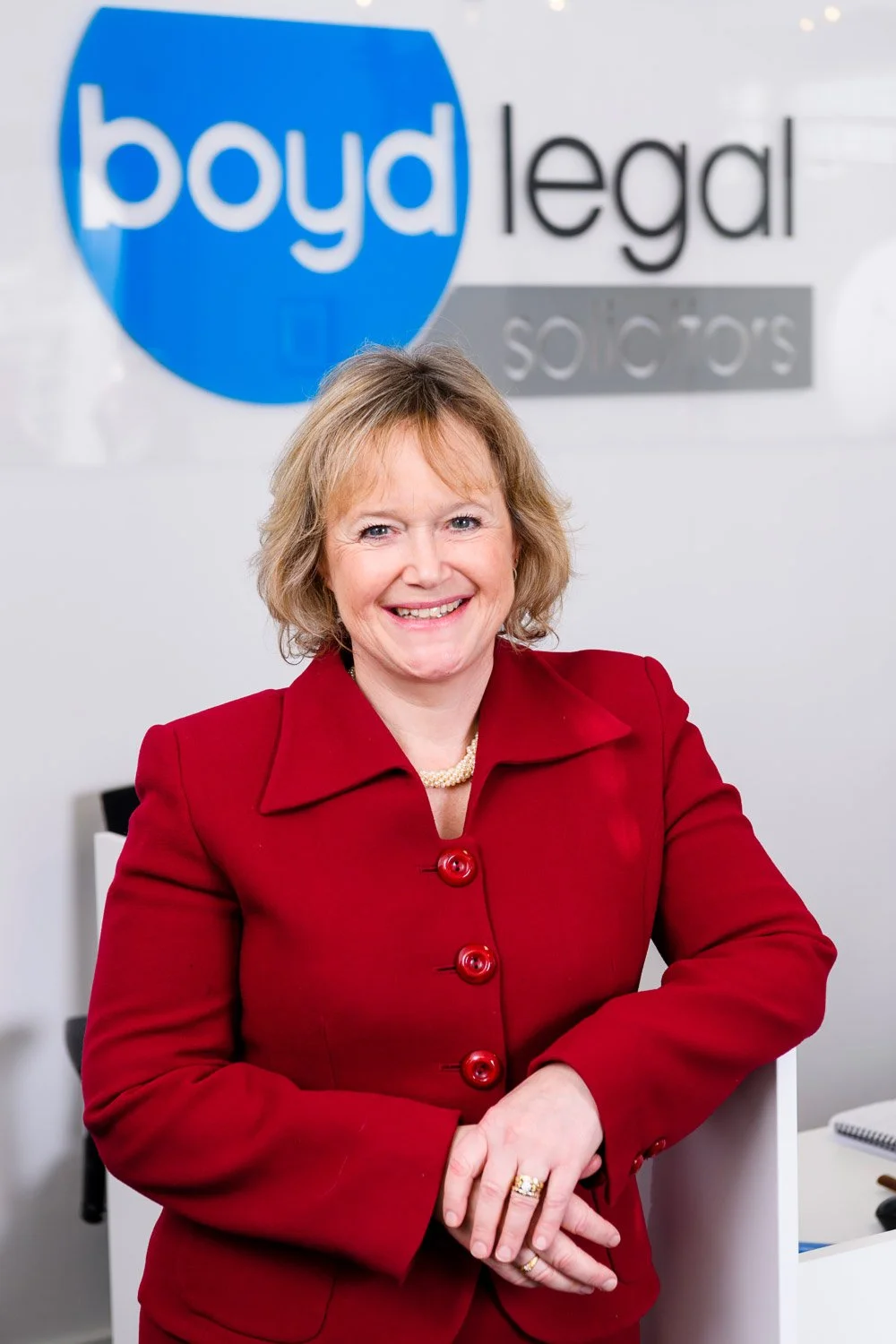 A smiling woman in a red blazer standing in an office with a Boyd legal sign in the background.