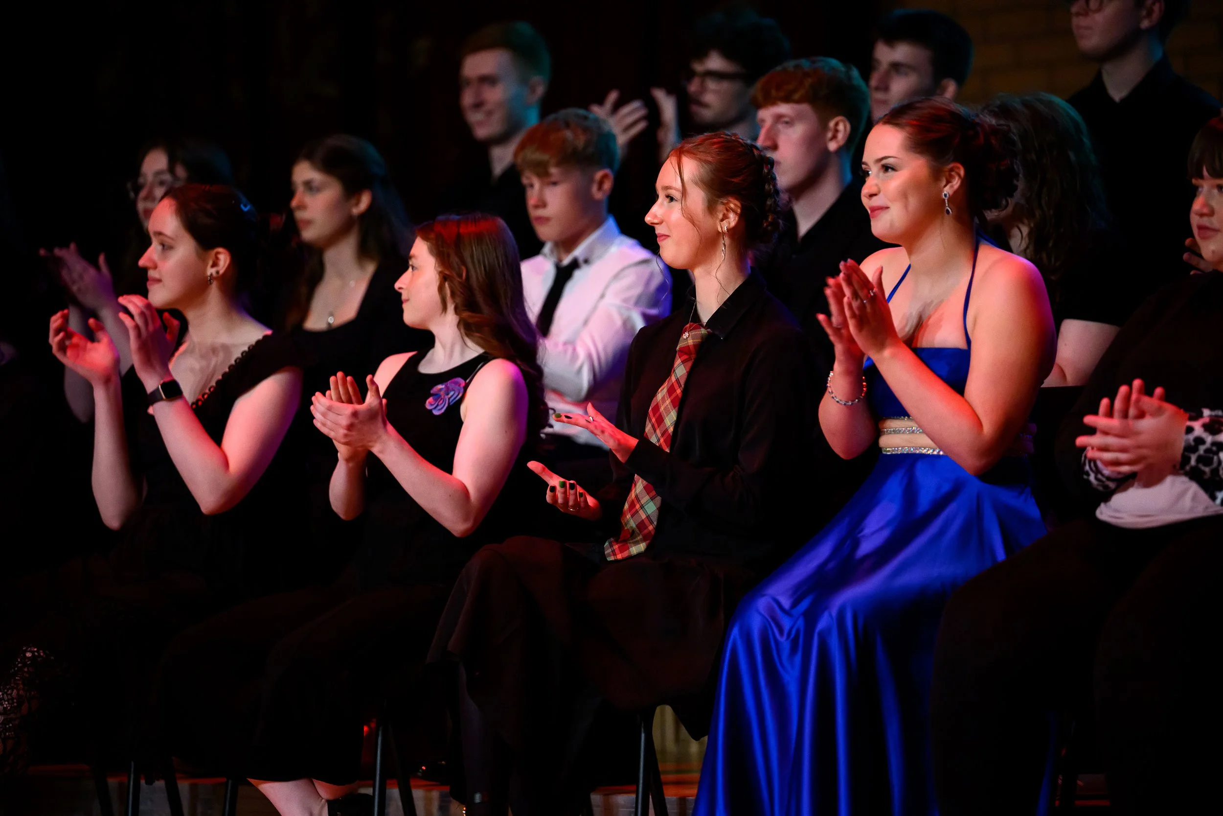 Audience members, mostly young women, applauding at an indoor event, with some dressed in formal attire.