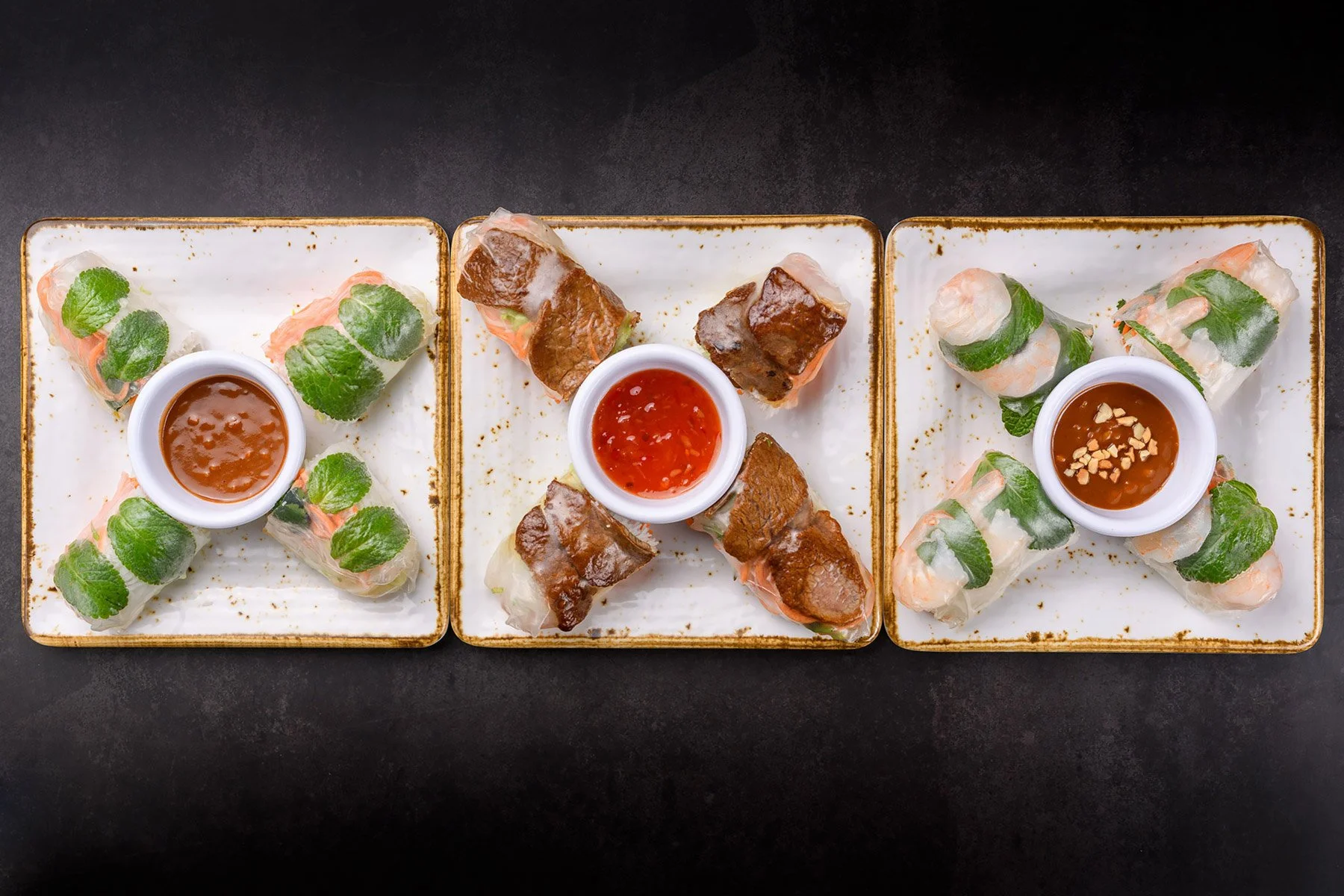 Three plates of Vietnamese spring rolls with dipping sauces on a black surface. The left plate has shrimp and herbs, the middle has grilled pork, and the right has shrimp and herbs. Each plate has a dipping sauce in a white bowl.
