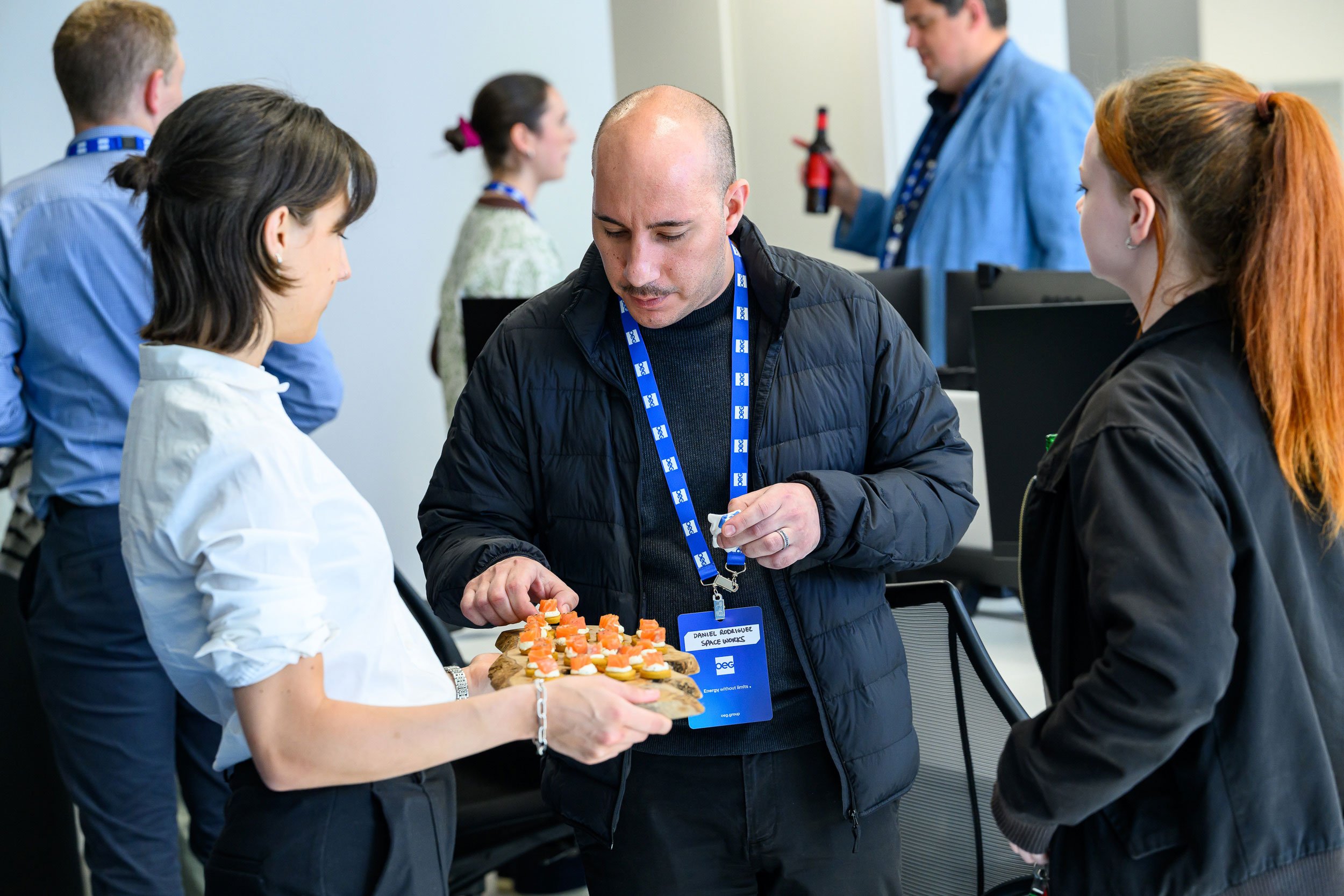 A man in a black jacket with a blue lanyard and name tag is holding a plate with small appetizers, talking to two women in black jackets, one with red hair and one with dark hair. Other people with blue lanyards are in the background at a professiona