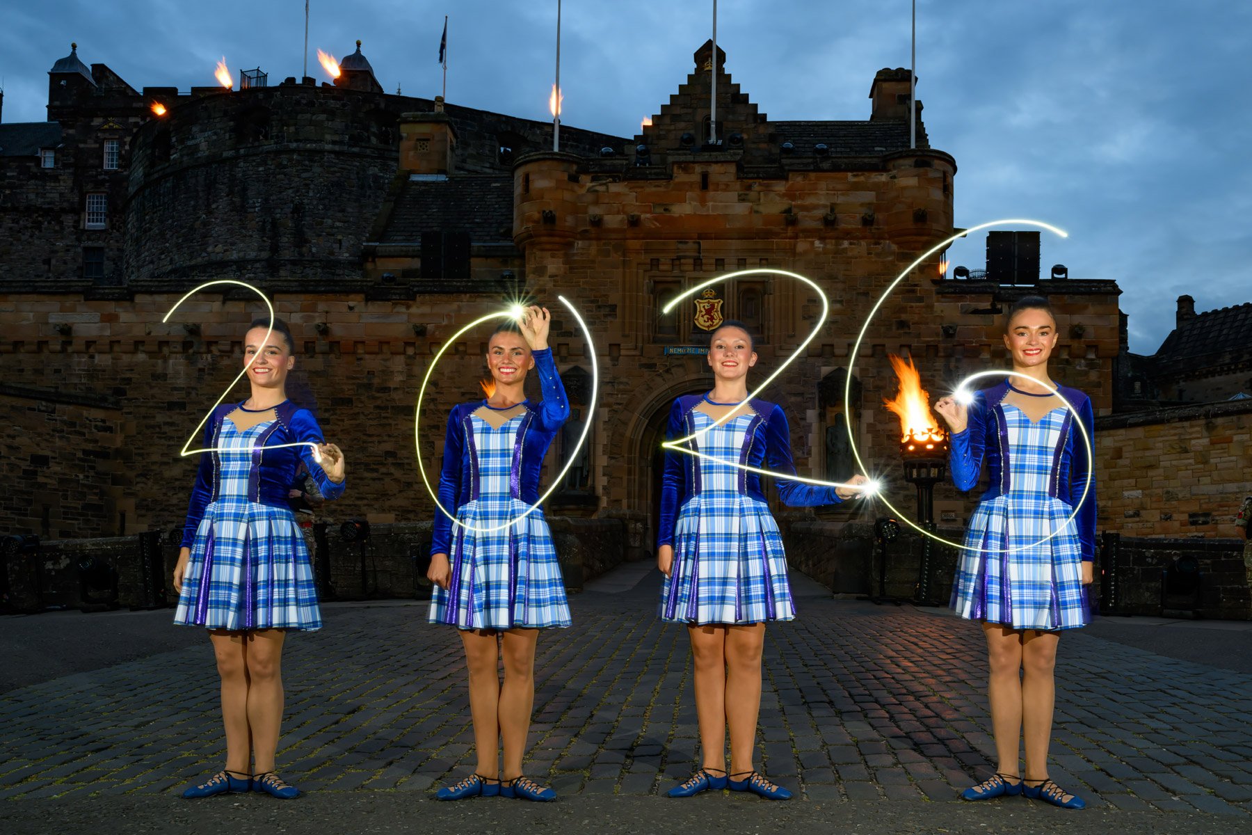 Four women dressed in traditional Scottish attire hold sparklers, forming the year 2023 with light trails in front of a historic castle at dusk.