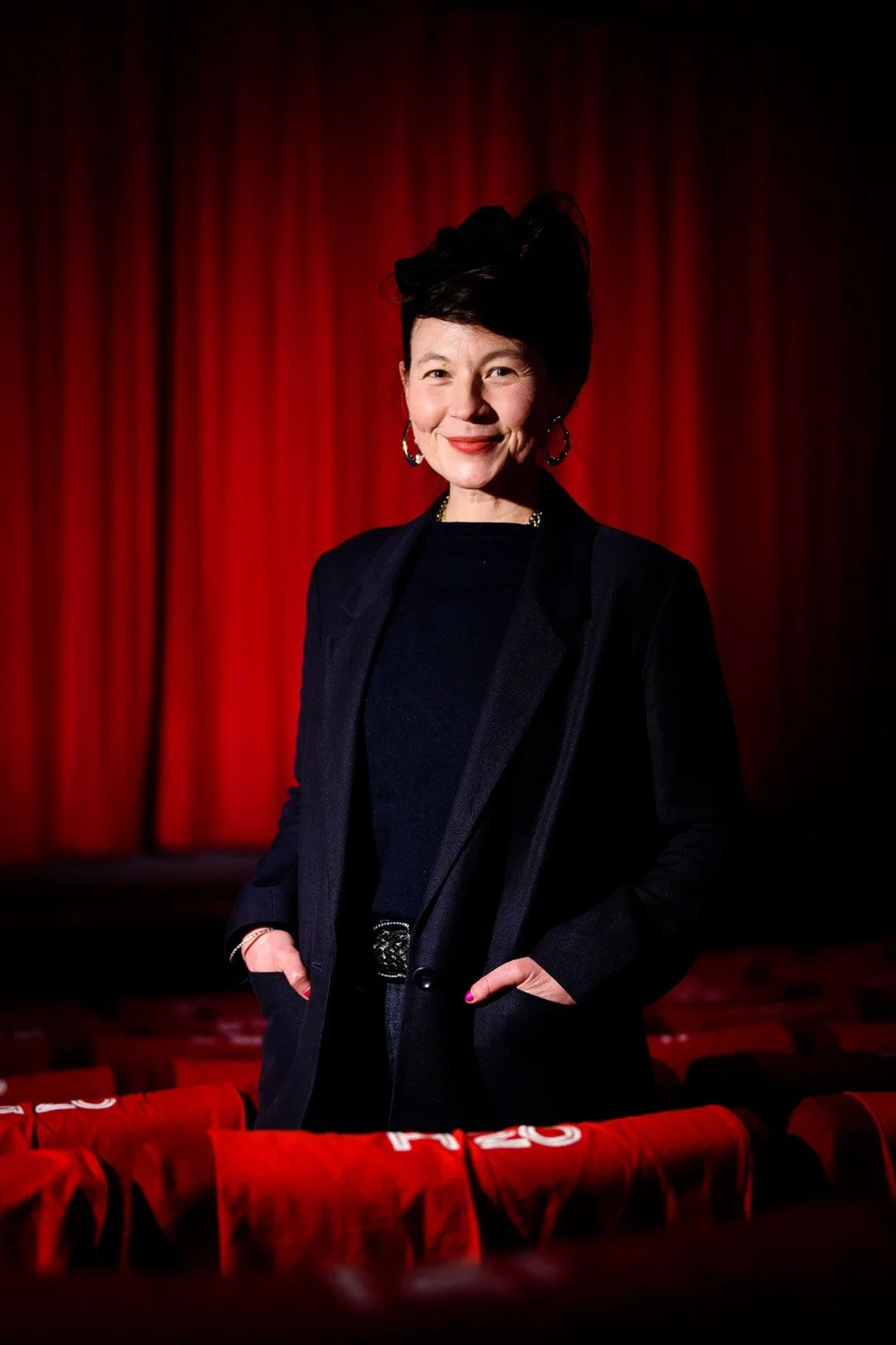 A woman standing in front of a red theater curtain, dressed in a black blazer and top, with earrings and a belt, smiling and posing for the camera.