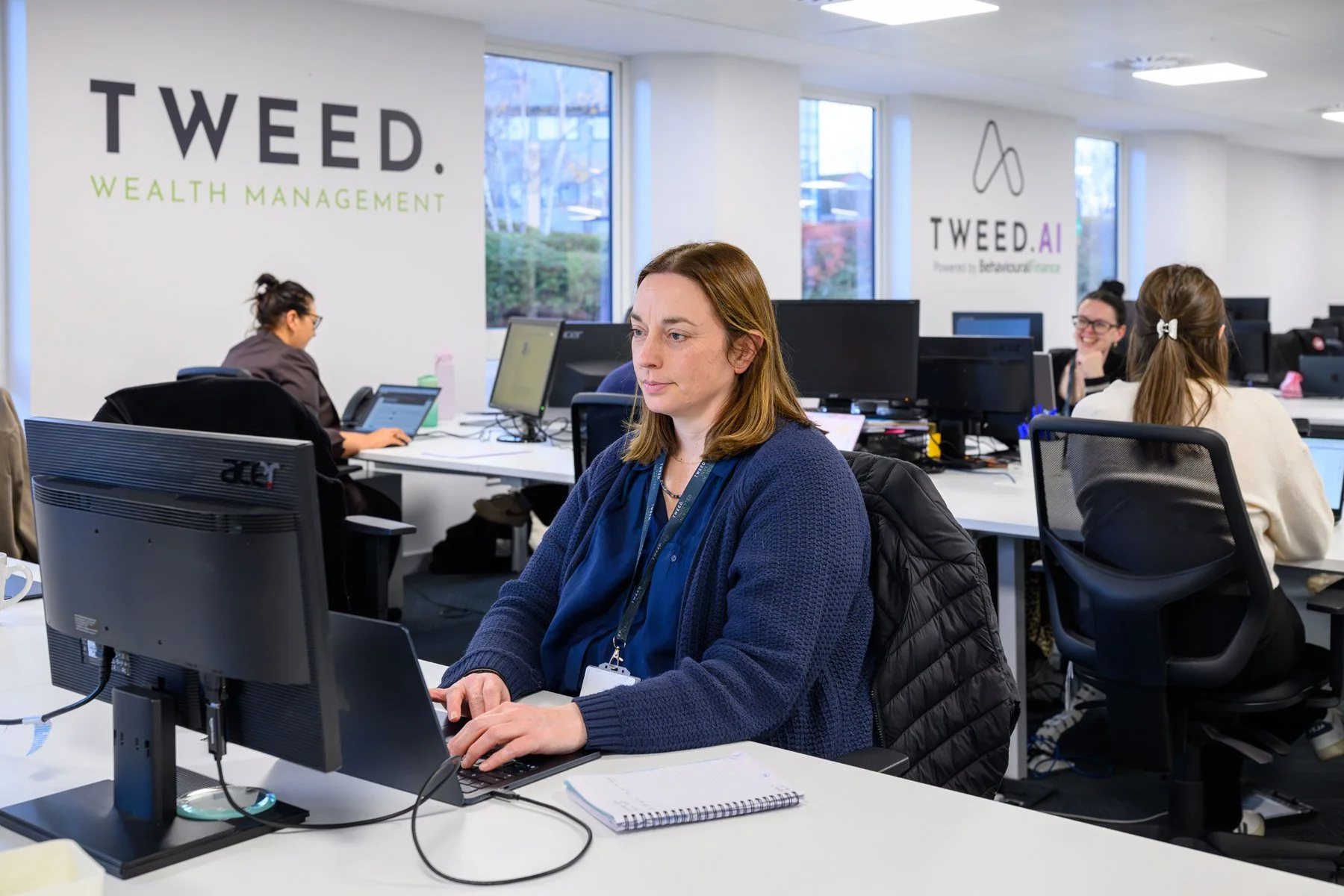 A woman working on a computer in an office with multiple desks and colleagues in the background, with logos on the wall reading 'TWEED. WEALTH MANAGEMENT' and 'TWEED.AI'.