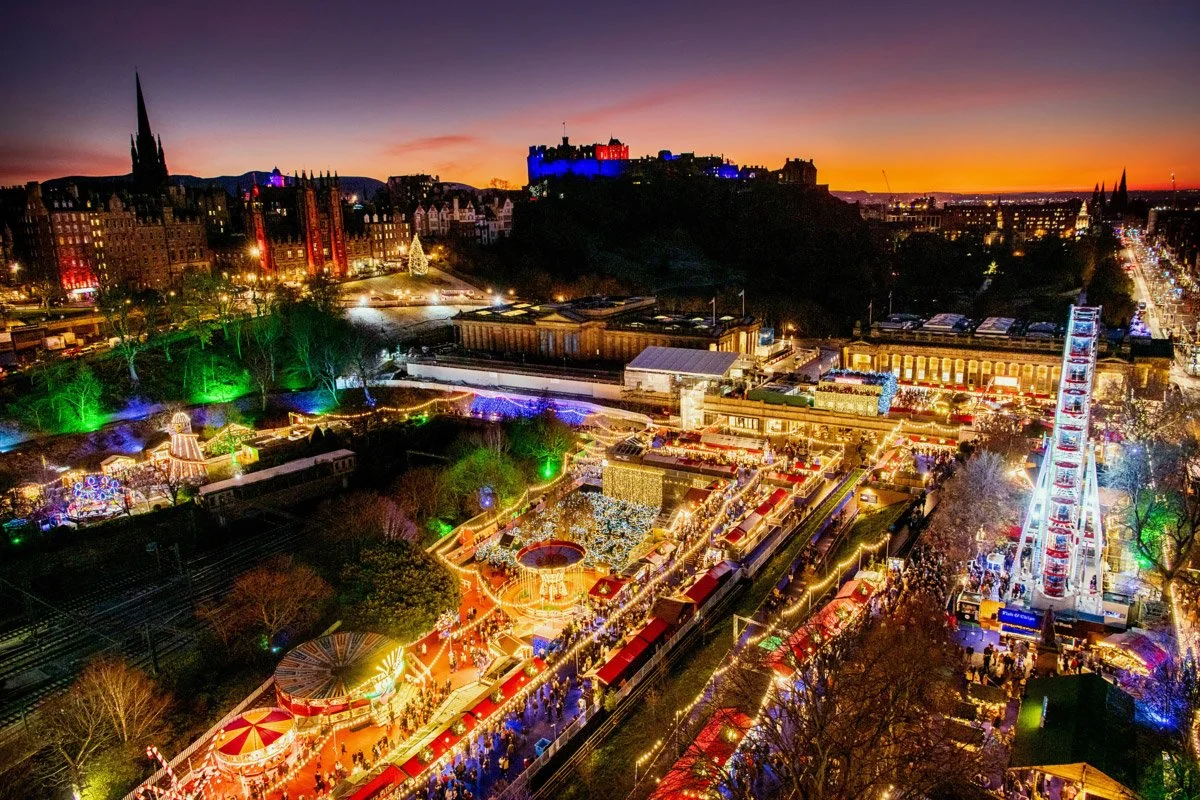 Nighttime view of a city park decorated with colorful holiday lights, with a large ferris wheel, amusement rides, and a crowd of people enjoying the festivities.