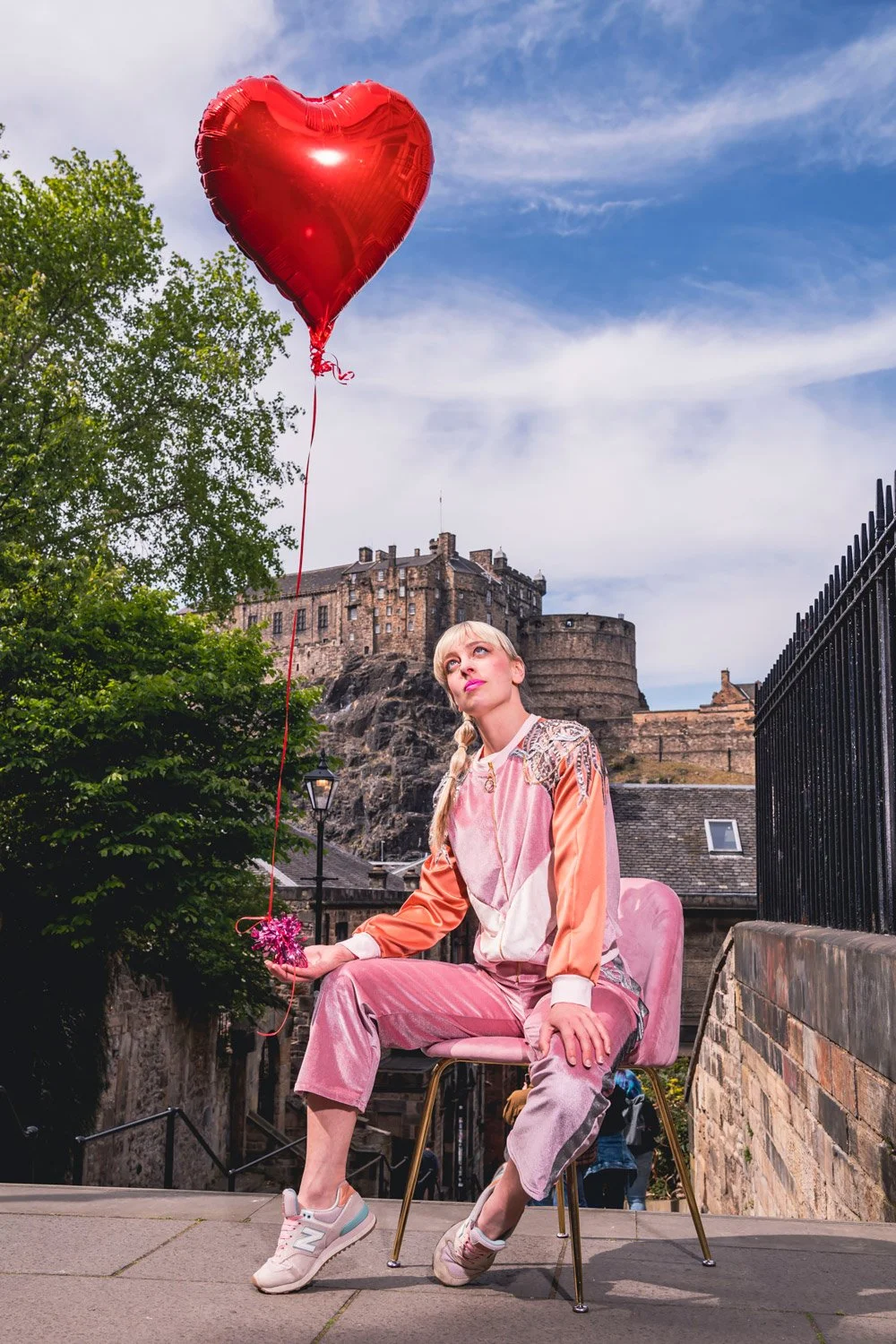 A woman sitting on a pink chair outdoors, holding a pink bouquet and a red heart-shaped balloon, with a historic castle and cloudy sky in the background.