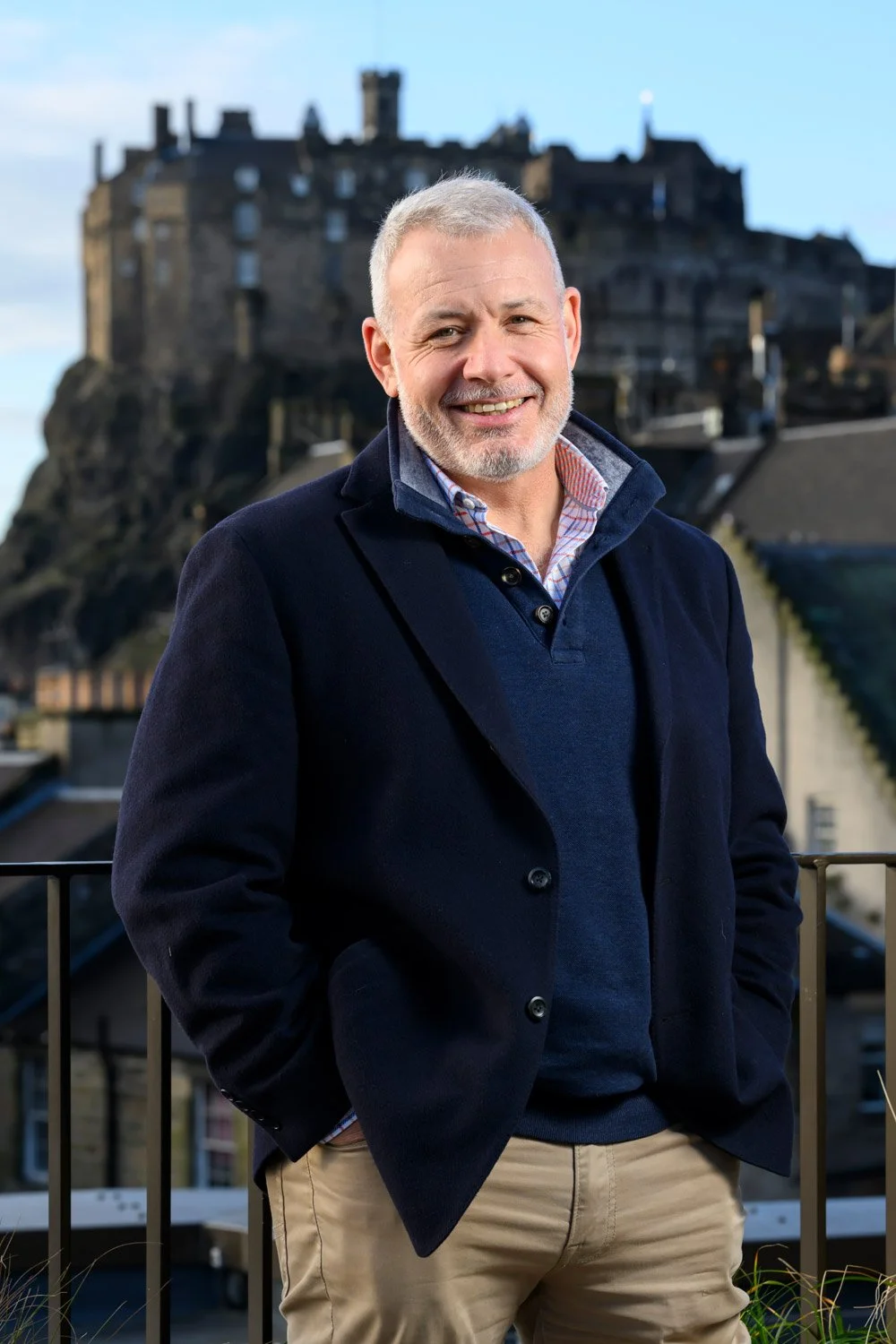 A middle-aged man with gray hair and a beard smiling, dressed in a dark blazer over a blue sweater and checkered shirt, standing outdoors with a castle on a rocky hill in the background.