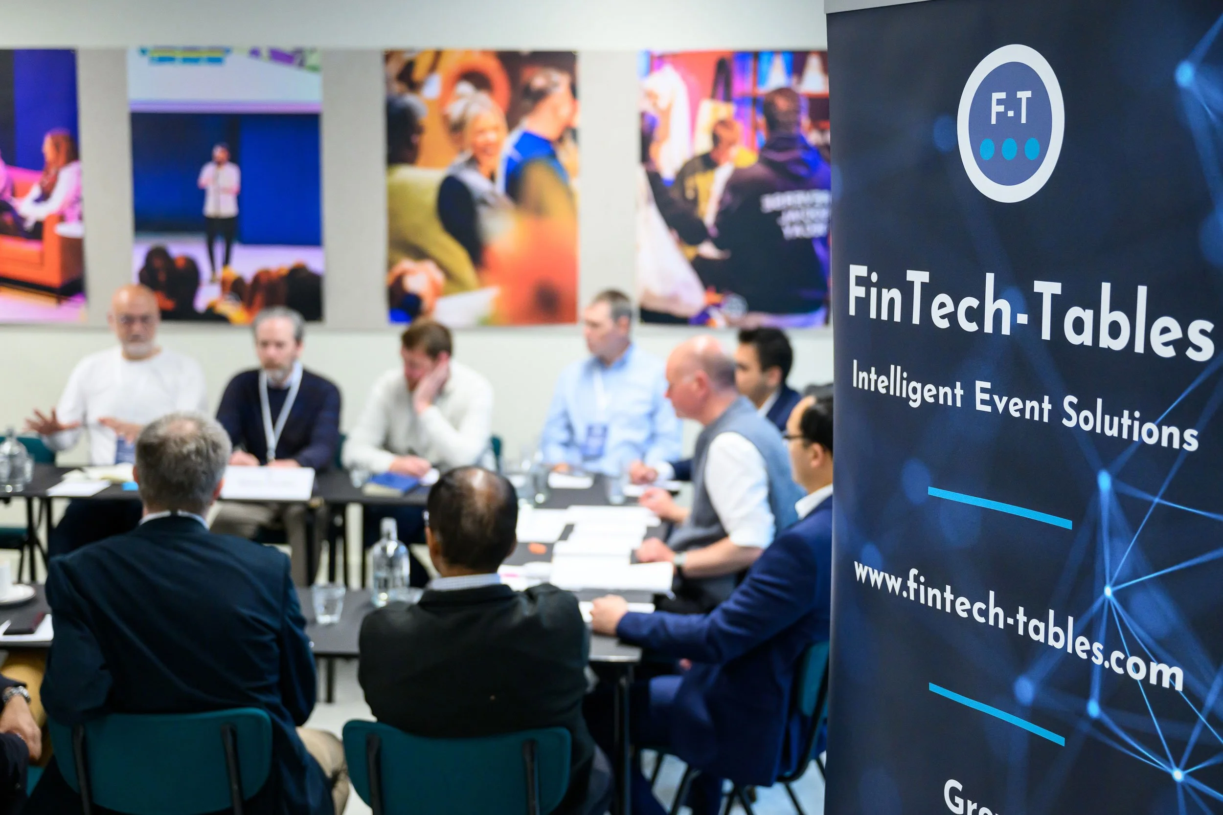A group of people seated around a conference table during a business meeting, with a banner on the right side that reads "FinTech-Tables, Intelligent Event Solutions, www.fintech-tables.com" and colorful artwork on the wall behind them.