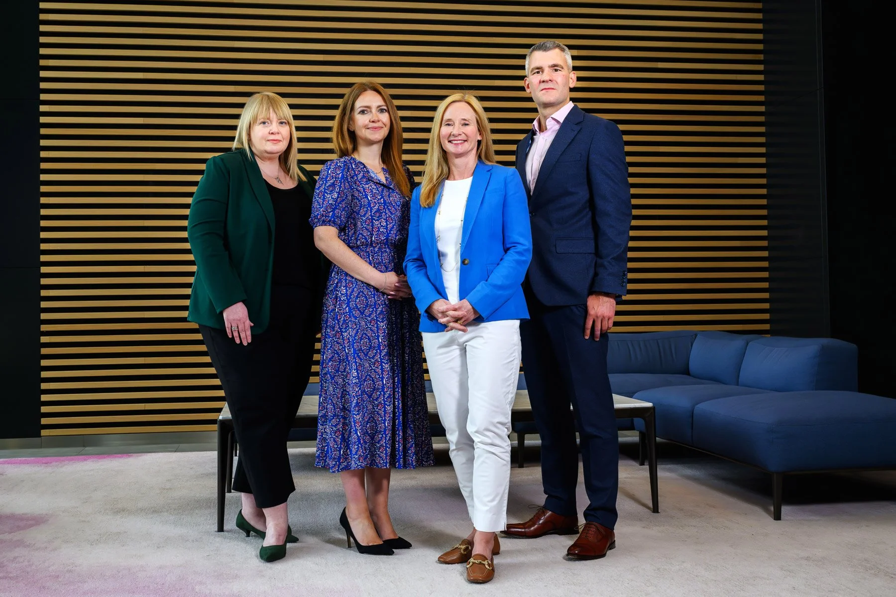 Four professionally dressed individuals, three women and one man, standing together in an indoor setting with wooden panel wall background and blue sofa.