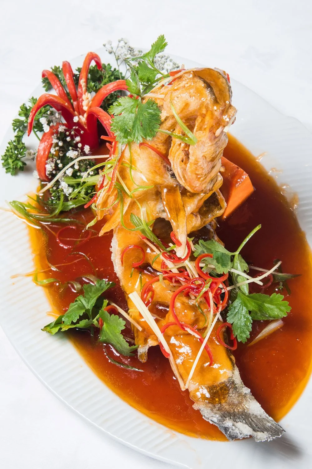A plate of cooked fish with sauce, garnished with fresh herbs, sliced chili peppers, and colorful vegetables.