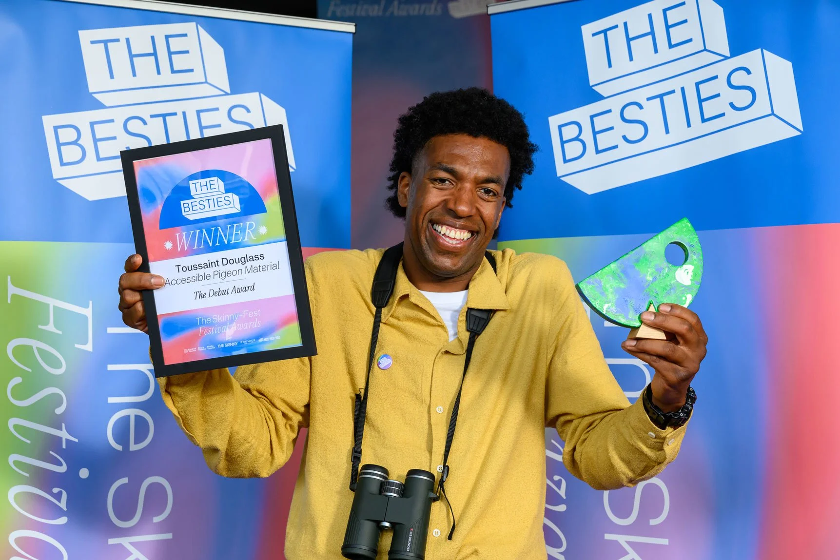 A man smiling and holding an award plaque and a colorful trophy at an event called 'The Besties.' The award plaque reads 'Toussaint Douglass, Accessible Pigeon Material, The Debut Award.' The man is standing in front of a backdrop with the event's lo