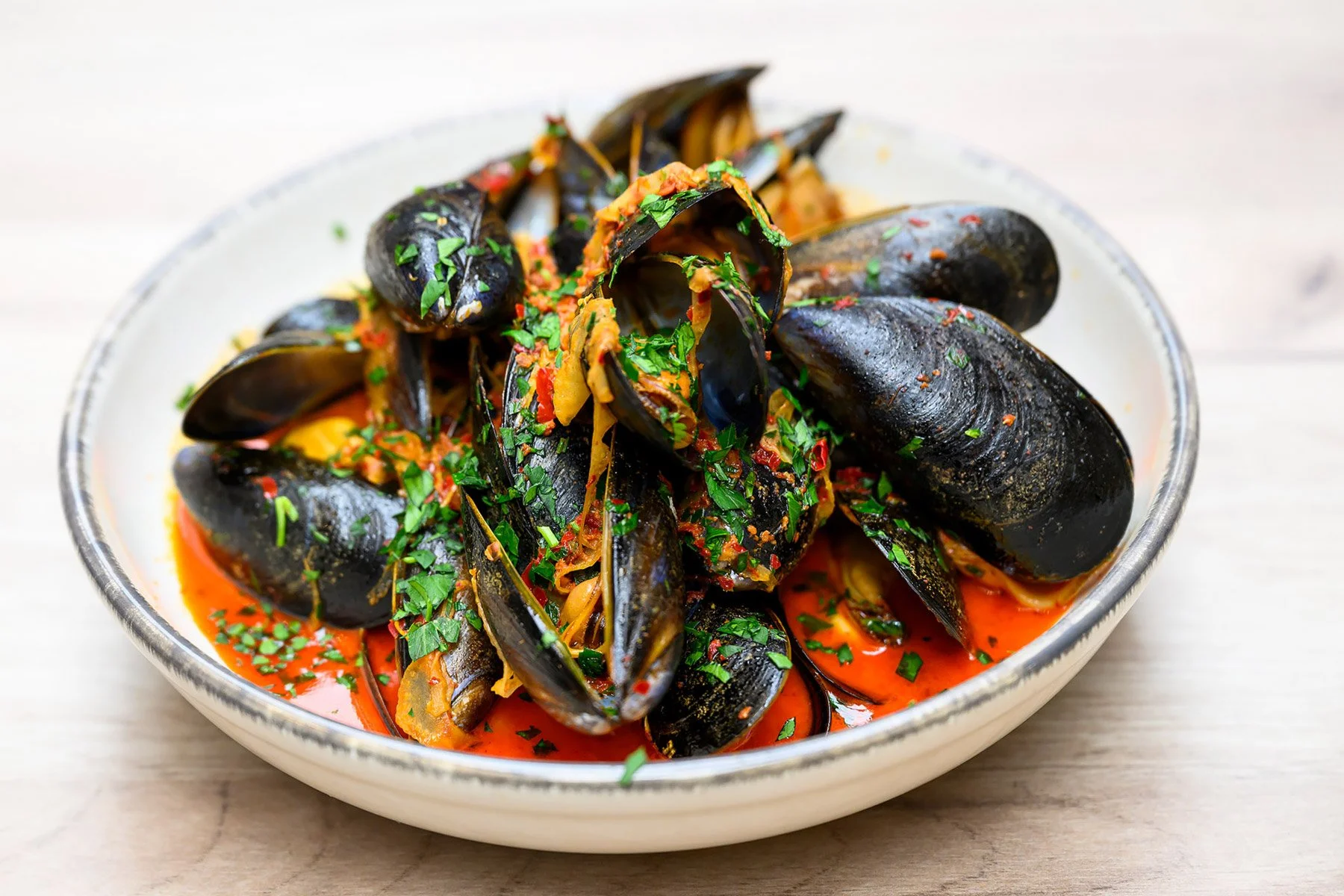 Bowl of mussels cooked in a spicy tomato sauce, garnished with chopped herbs, served in a white bowl on a light wooden surface.