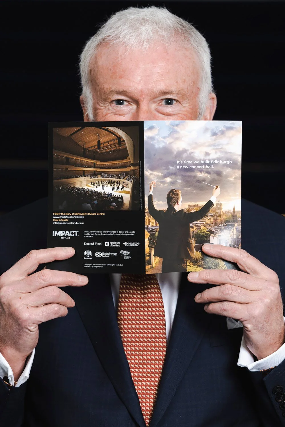 Man in suit holding a promotional brochure in front of his face, revealing only his eyes, promoting a new concert hall in Edinburgh with images of an interior concert hall and an outdoor city view.