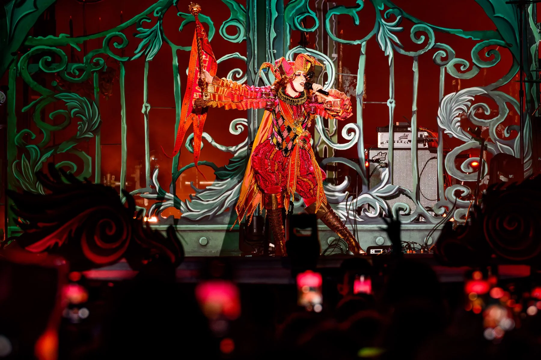 Performer dressed in vibrant, elaborate red and orange costume with jester-like hat, performing on stage with ornate green and white backdrop.