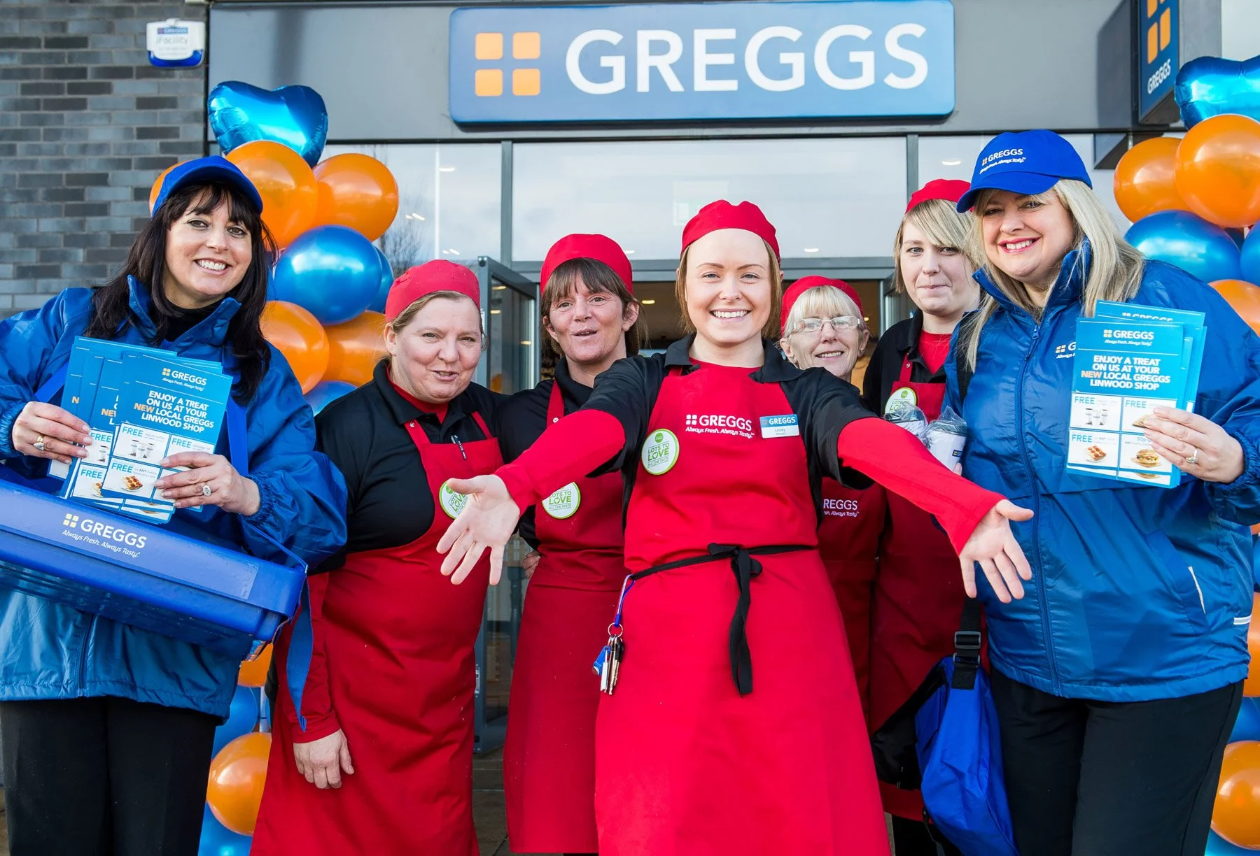 Group of seven smiling store employees standing outside Greggs bakery, some wearing red aprons and others in blue jackets and hats, with balloons and promotional pamphlets.