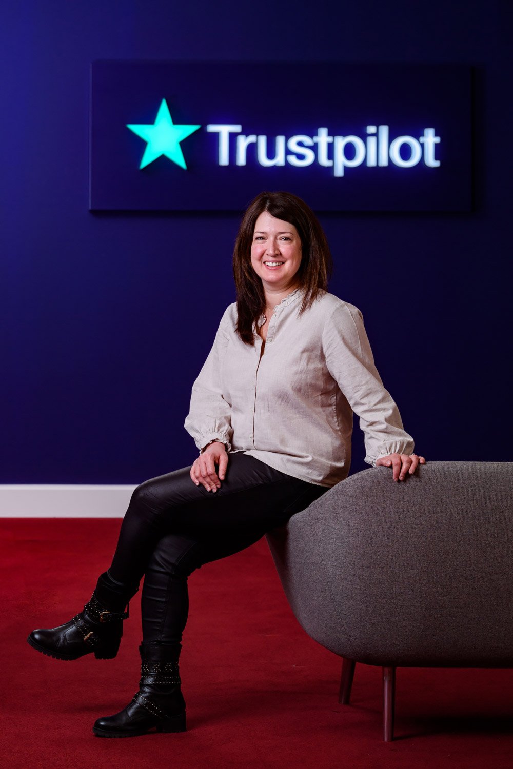 A woman sitting on a gray chair in front of a dark blue wall with a neon Trustpilot sign.