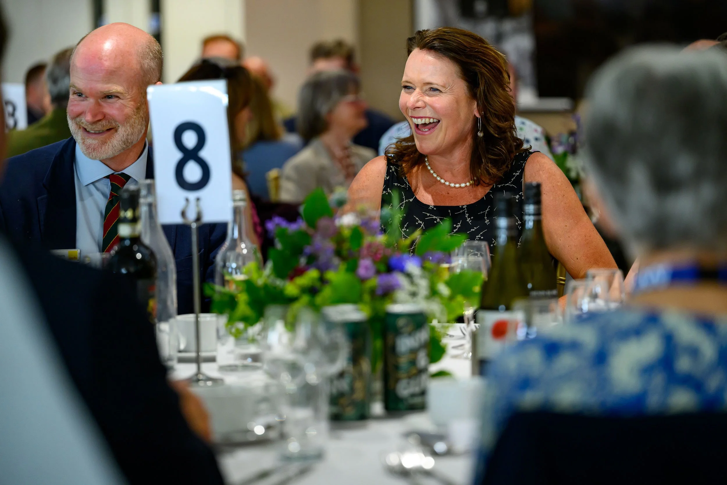 People attending a formal event or banquet, seated around a table with floral centerpieces and bottles, laughing and enjoying themselves.